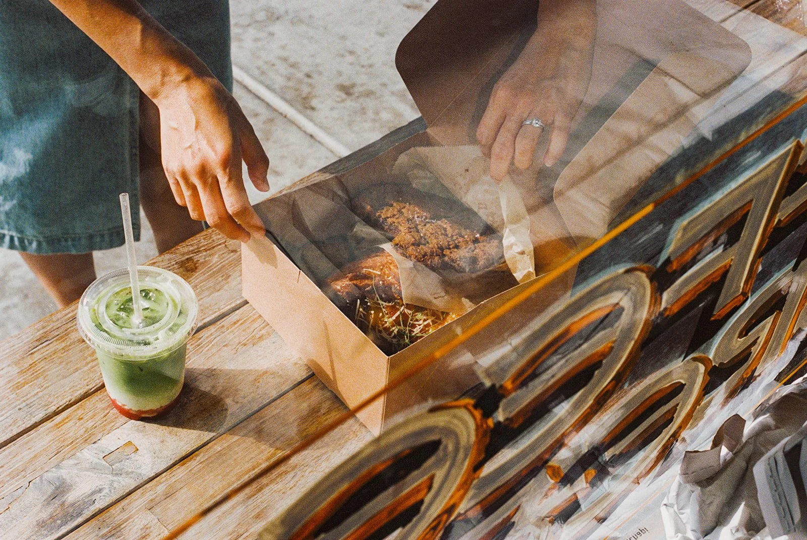 A person reaching into a paper takeout box of fried chicken on a wooden table, with a green drink in a plastic cup nearby.