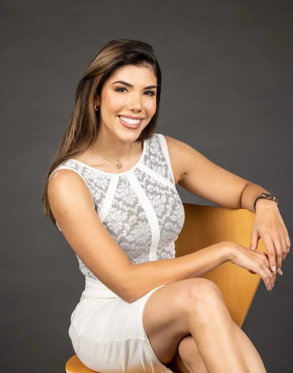 Dr. Maria Bernal-Riera with long brown hair, smiling, wearing a sleeveless white dress with lace details, sitting on a wooden chair against a gray background.