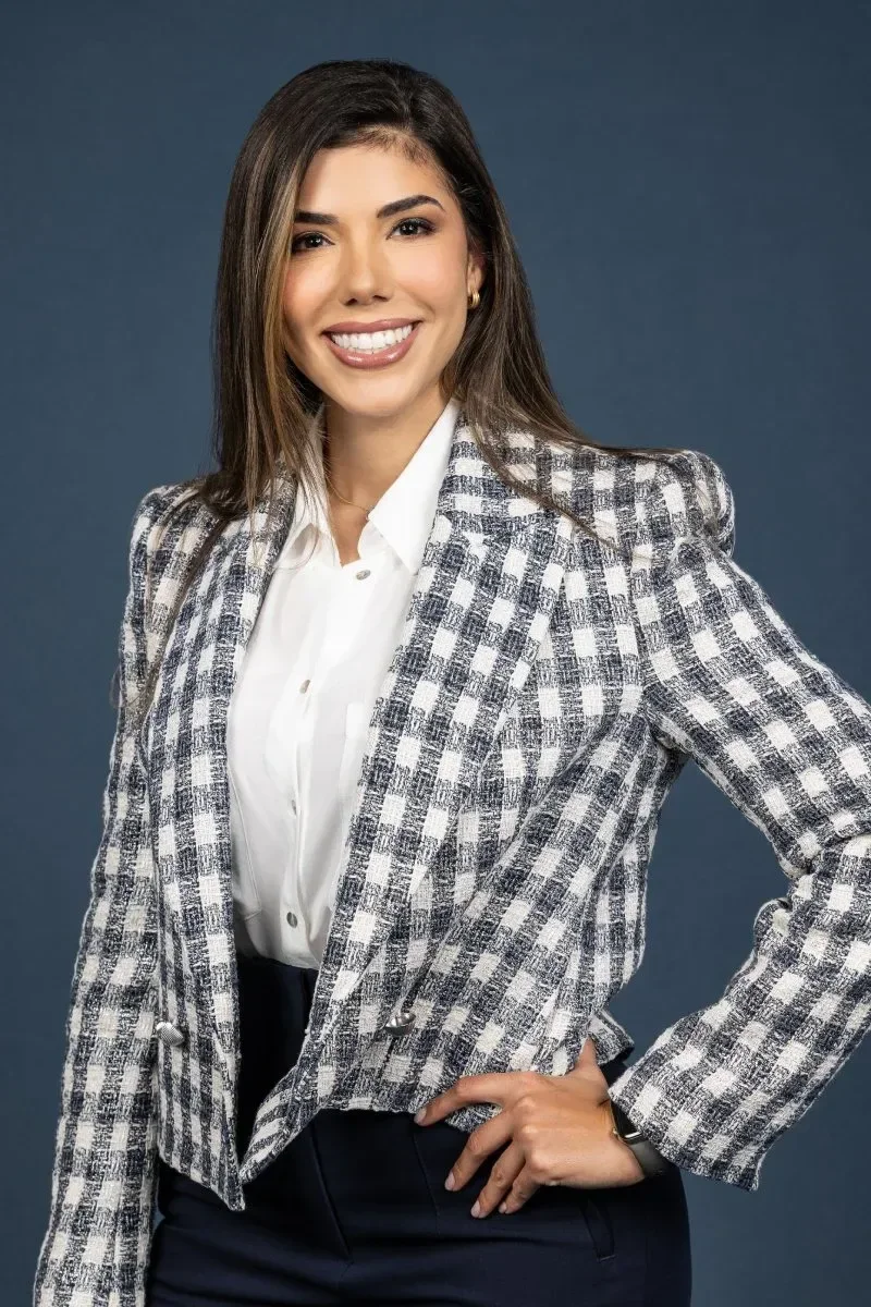 Dr. Maria Bernal-Riera with shoulder-length brown hair, wearing a plaid blazer over a white blouse and dark pants, standing against a solid blue background.
