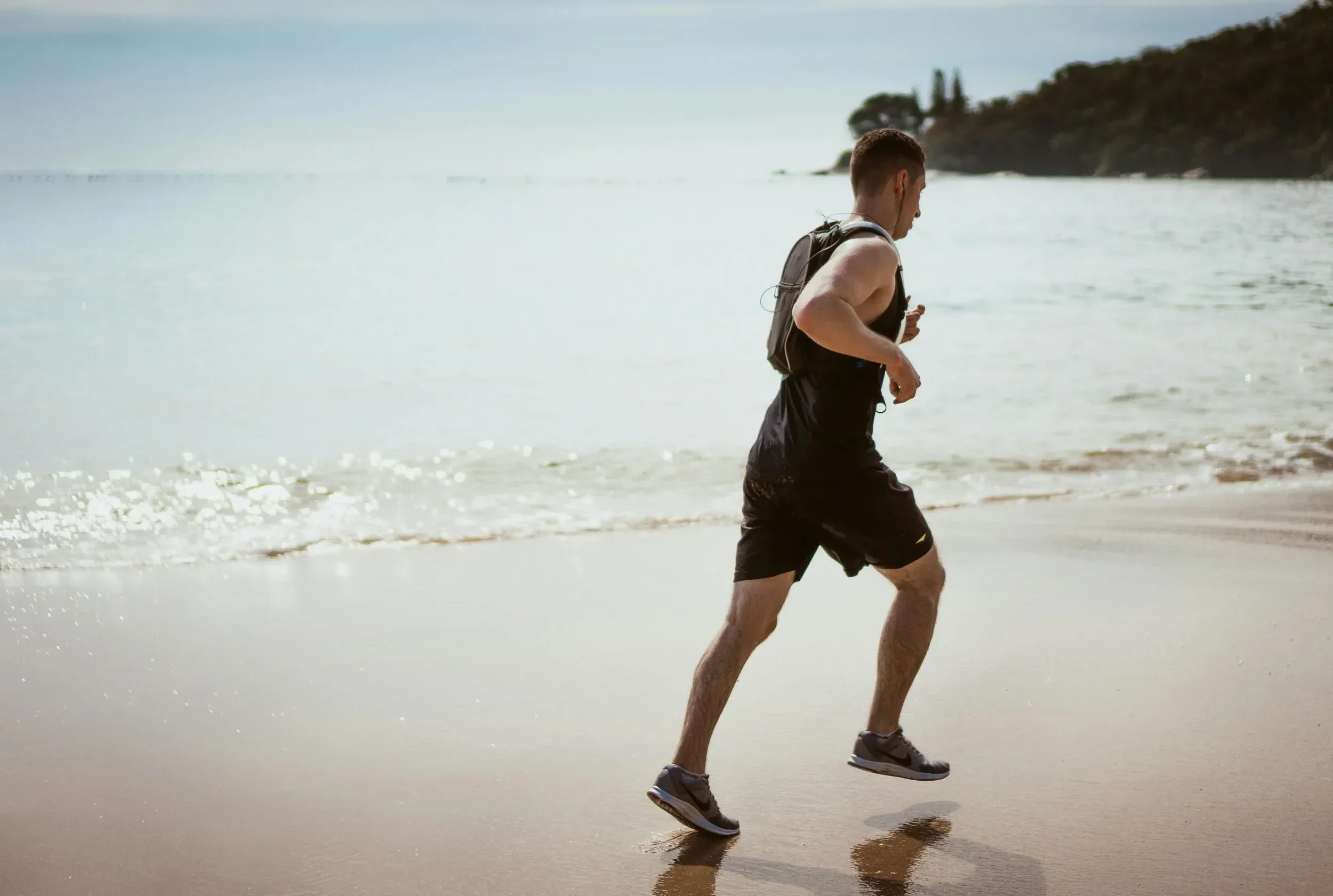A man running on a sandy beach near the ocean with a backpack.