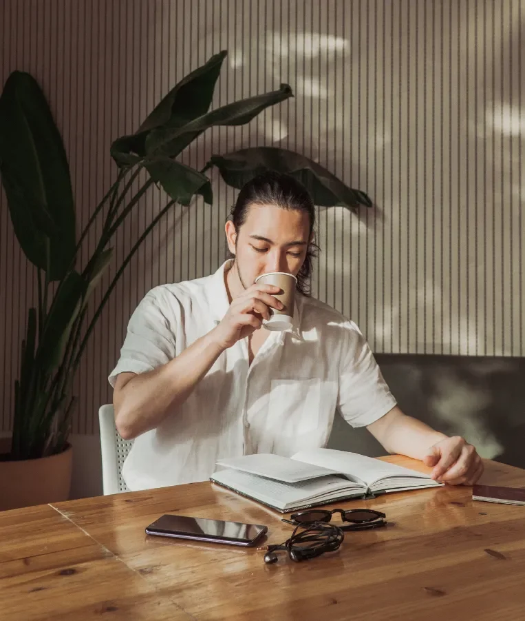 A person sitting at a wooden table, drinking coffee from a paper cup, reading a notebook, with a smartphone and glasses on the table, and large green plants behind them.