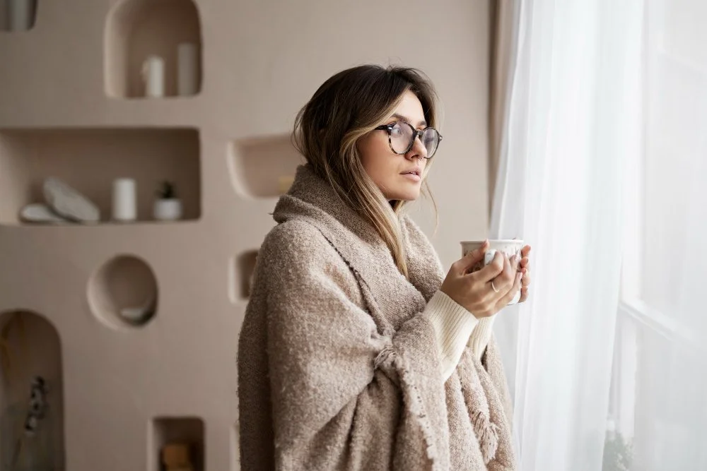 Woman wrapped in a knit shawl holding a warm mug by a window