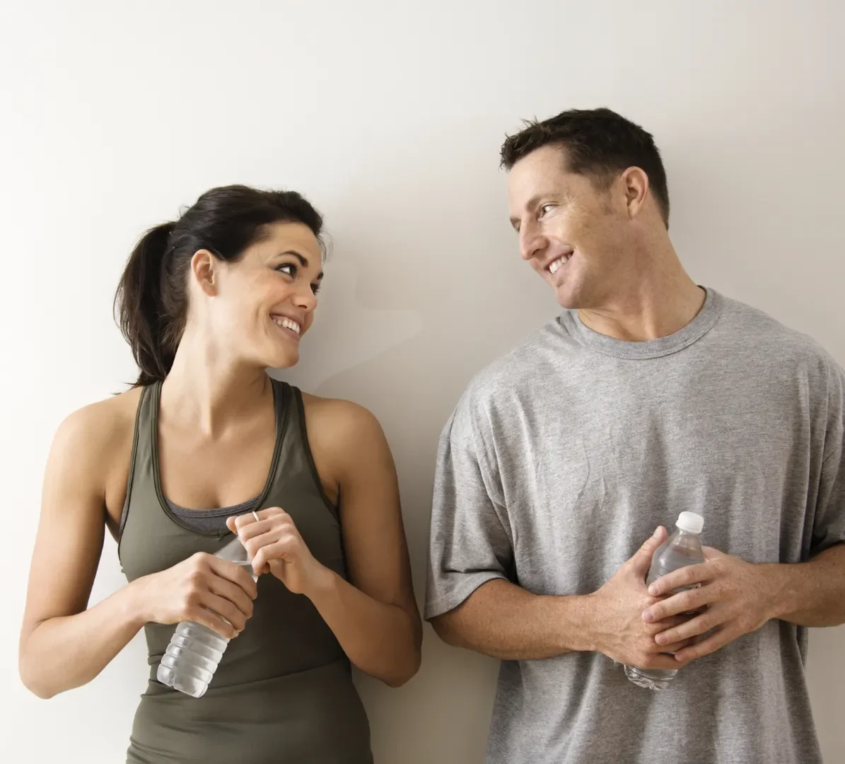 A woman and man smiling at each other while holding water bottles, casually dressed, standing against a plain white wall.