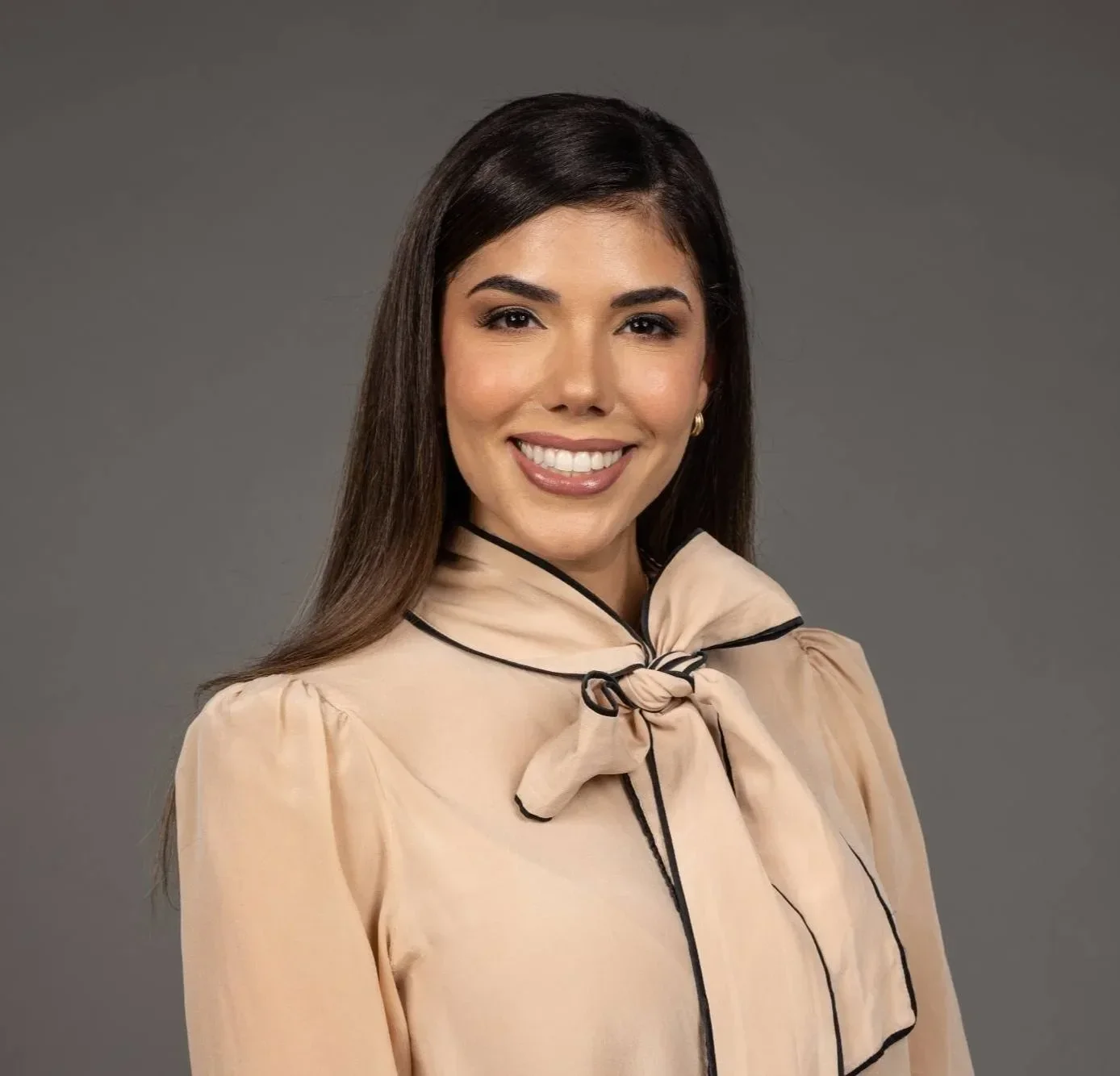 Dr. Maria Bernal-Riera with long brown hair, smiling, wearing a beige blouse with a bow tie at the collar, against a gray background.