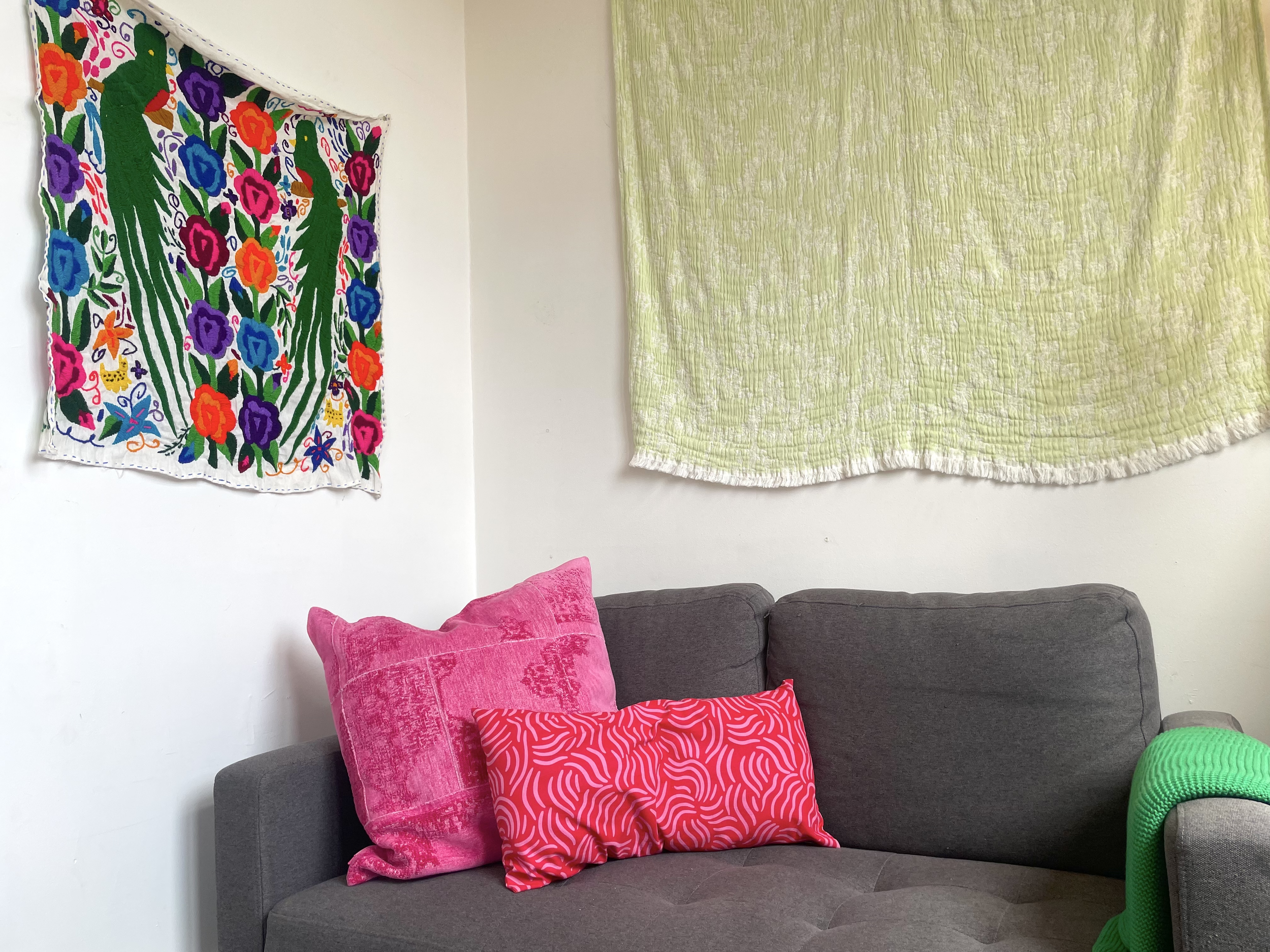 A gray sofa with three decorative pillows in pink, red and orange patterns. Two tapestries hang on the white wall above the sofa, one with colorful floral embroidery and the other in a light green textured fabric. A small green knitted object is partially visible in the bottom right corner.