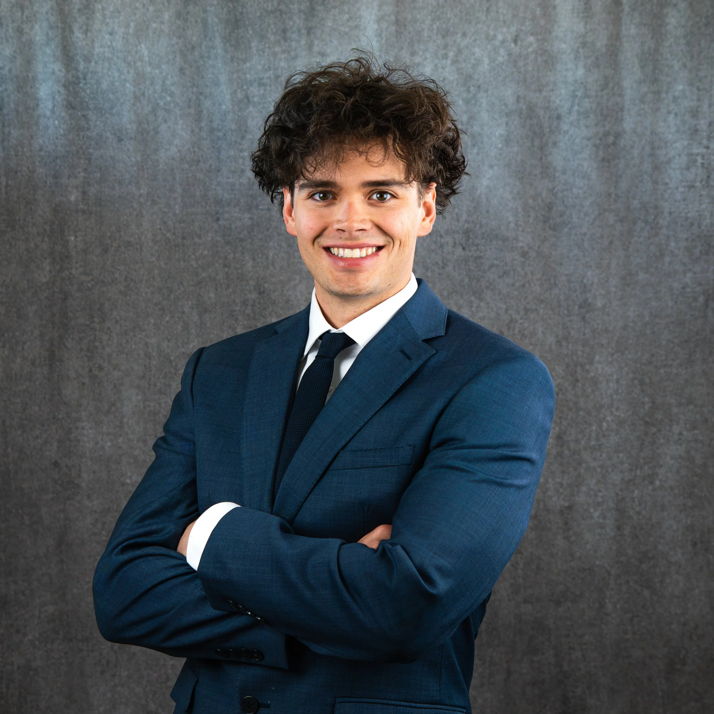 A young man with curly dark hair, wearing a navy blue suit and white shirt, smiling with arms crossed, standing against a gray background.