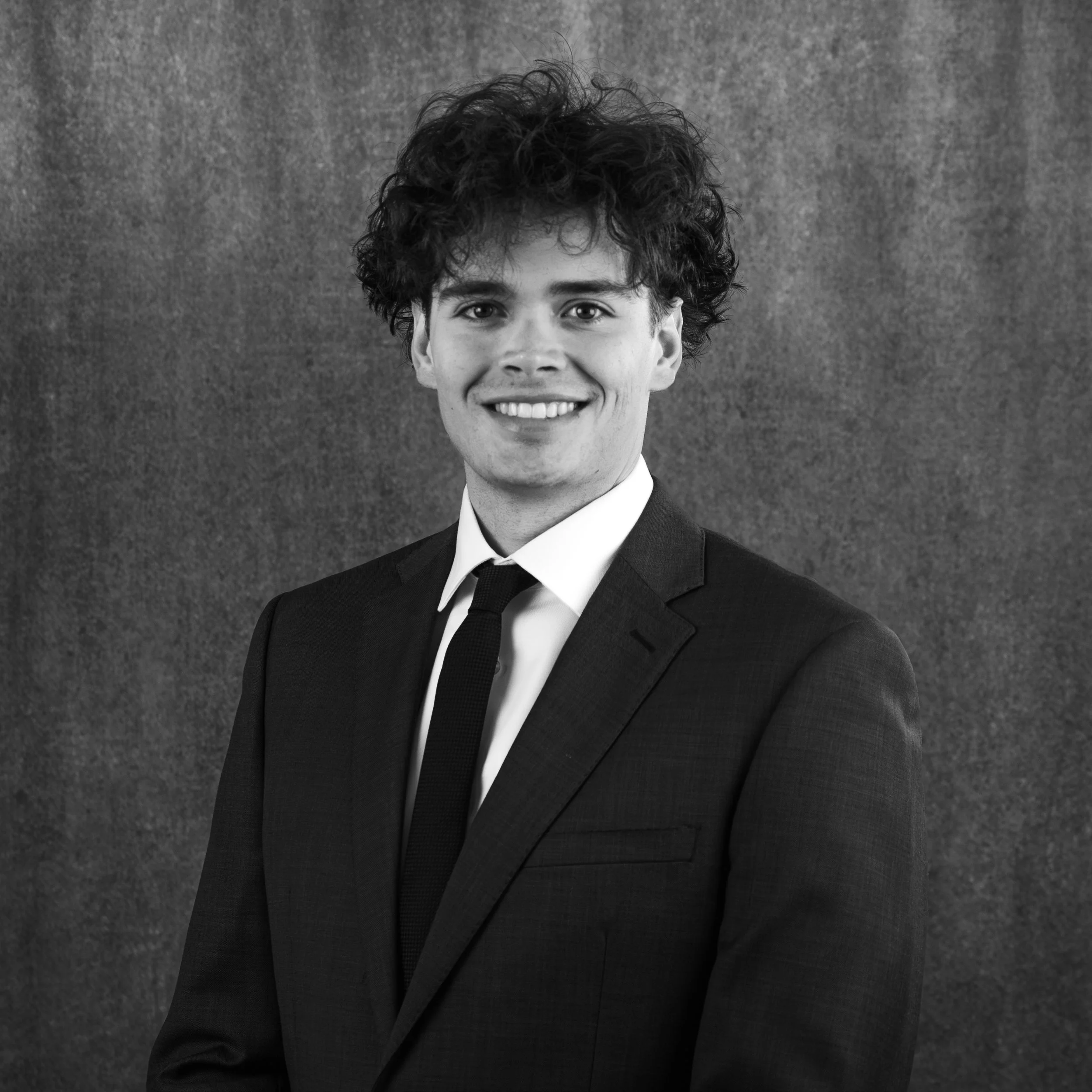 A young man with curly hair, smiling, wearing a suit and tie, against a textured gray background.