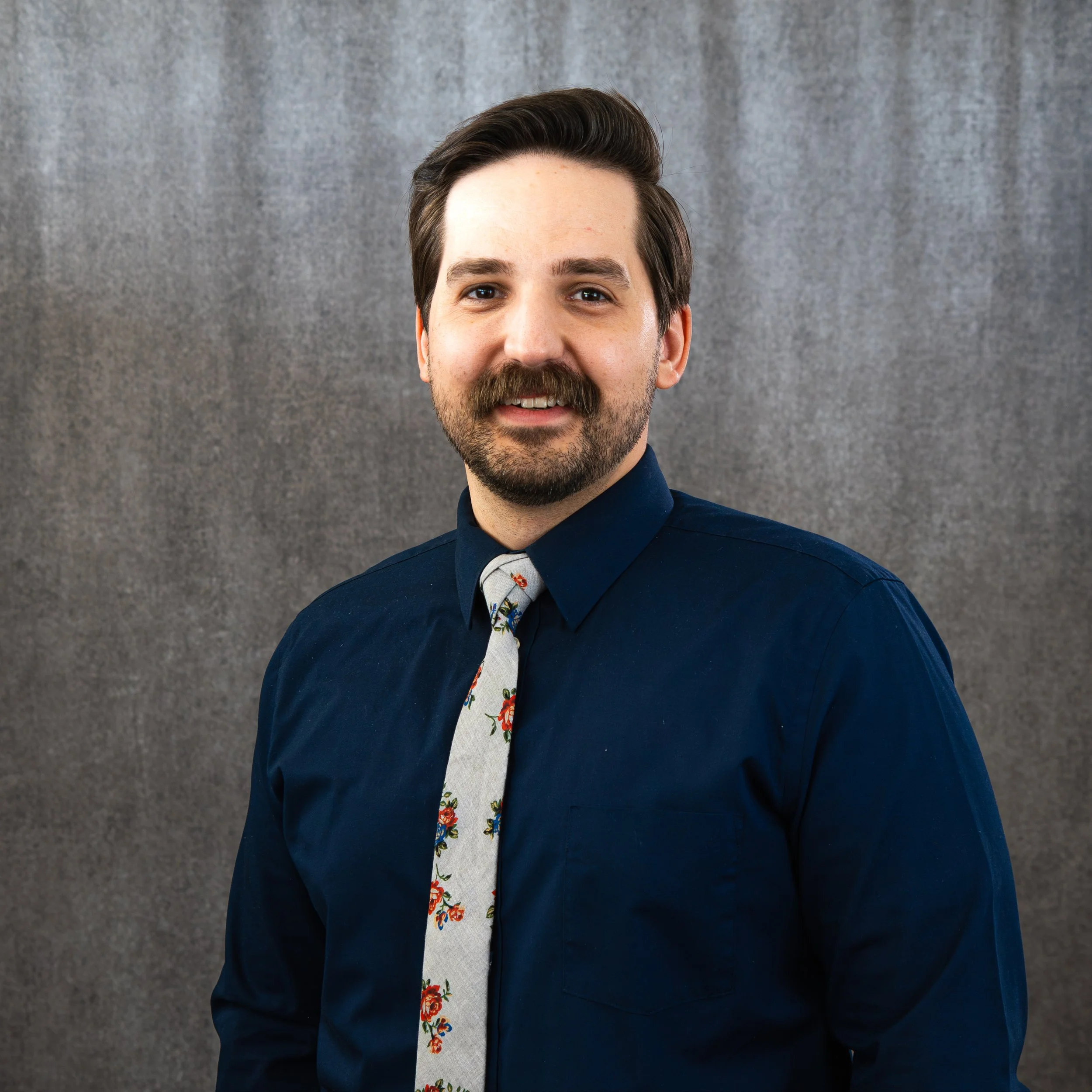 Close-up of a smiling man with dark hair and a beard, wearing a navy blue blazer and a light-colored shirt with a floral pattern, standing against a gray textured background.