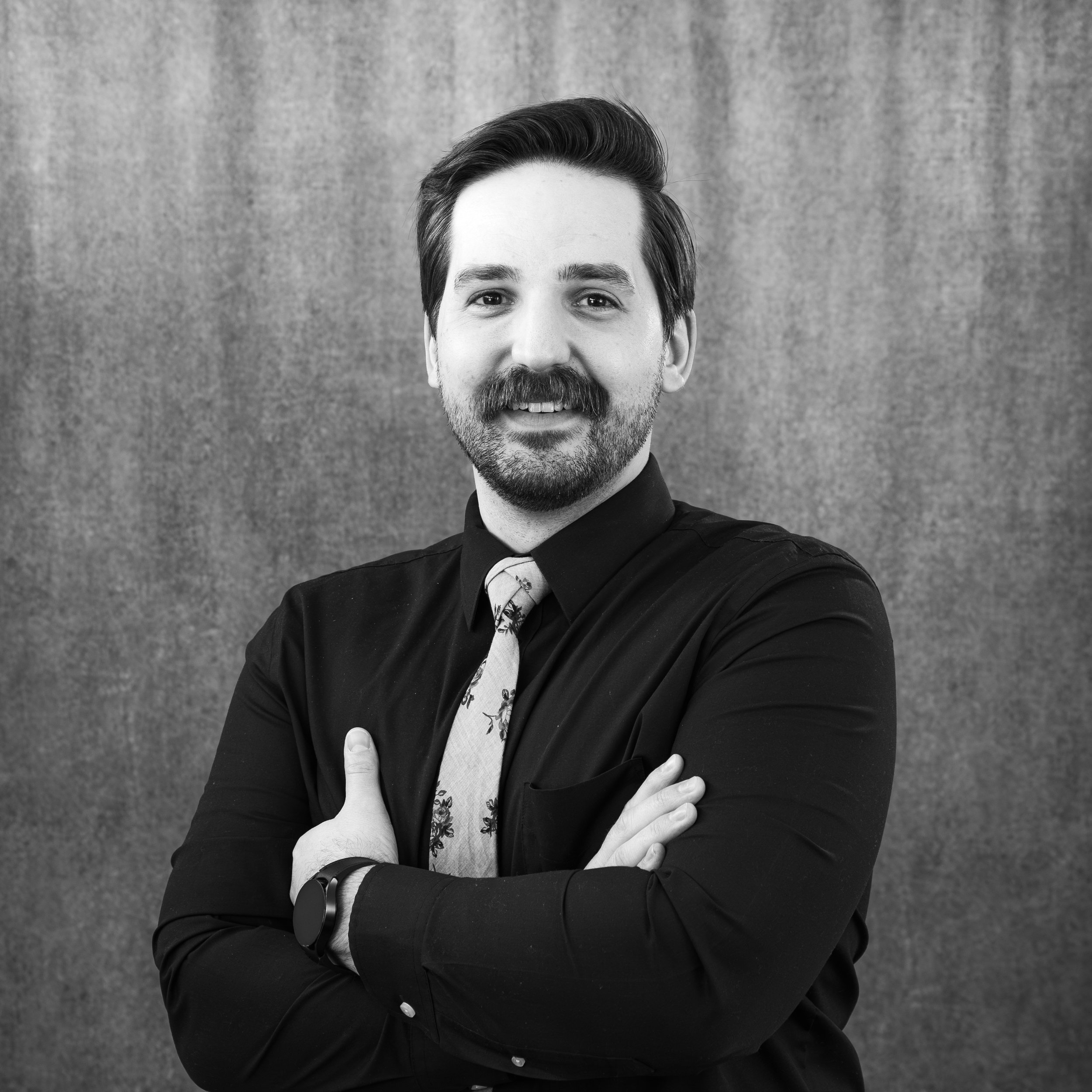 Black and white portrait of a young man with styled hair and a beard, wearing a black suit and a patterned tie, standing against a textured background.