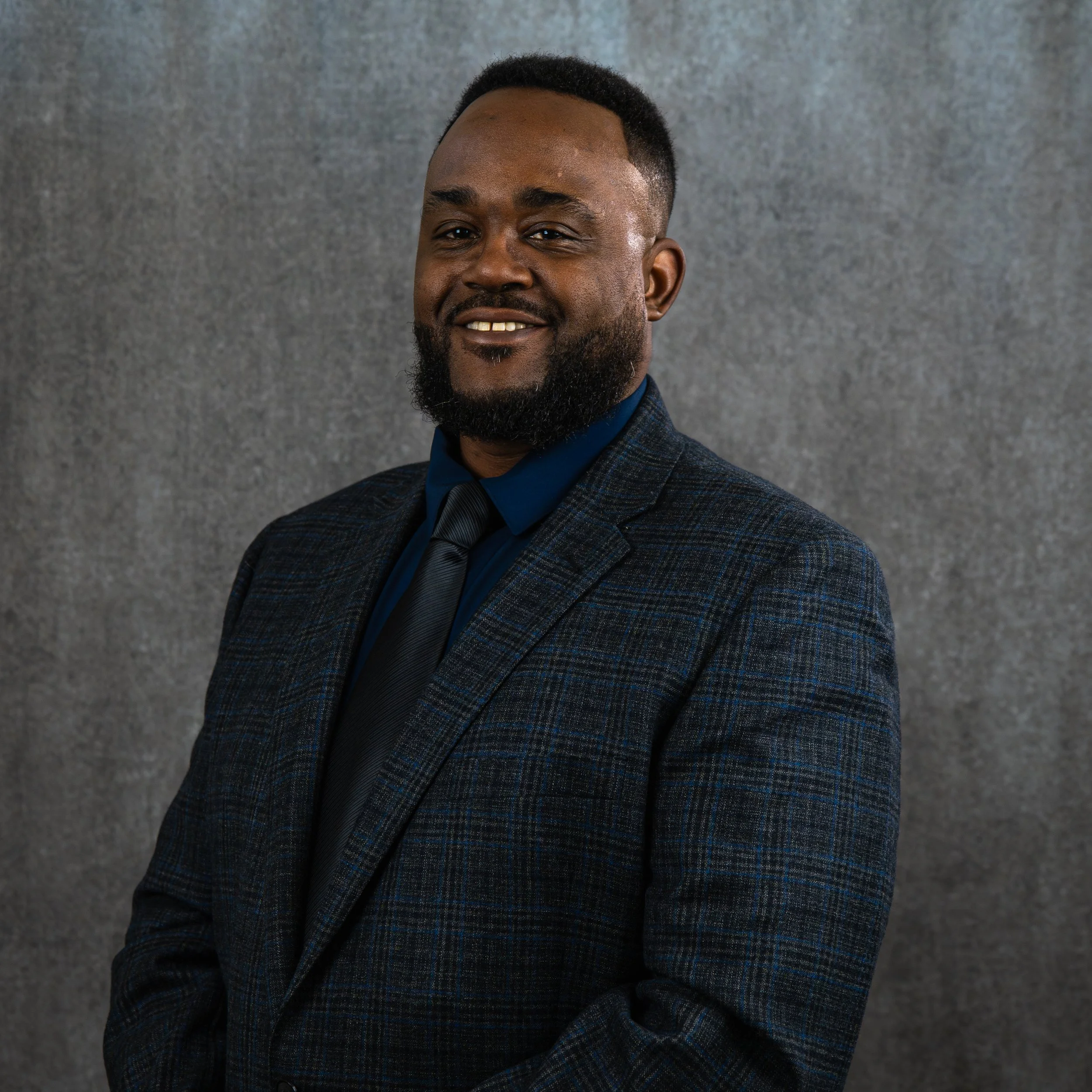 A portrait of a smiling Black man with a beard and short curly hair, wearing a dark checkered blazer and a blue shirt, standing against a textured gray background.