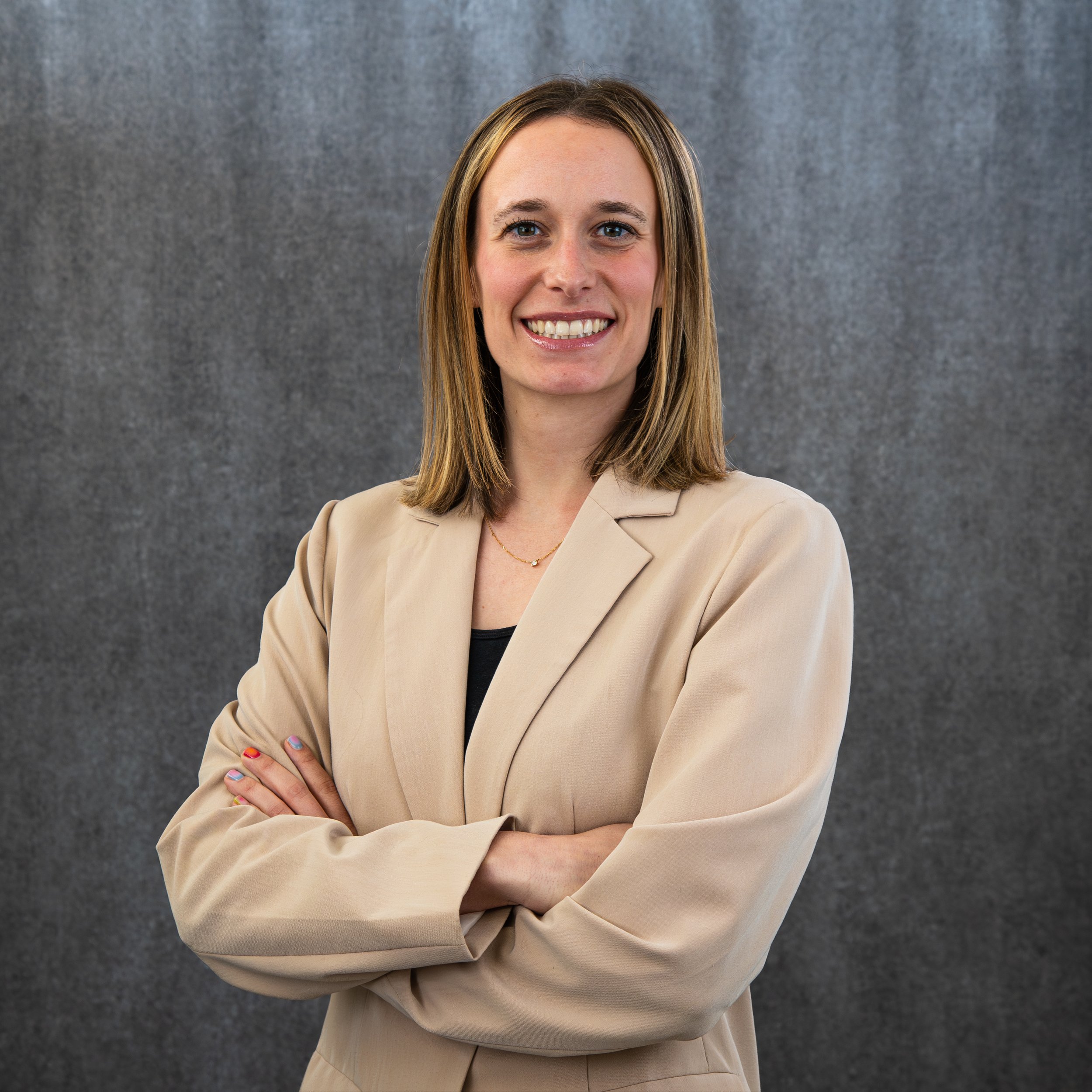 A woman with straight, shoulder-length blonde hair, blue eyes, and a friendly smile, standing against a gray, textured background. She is wearing a beige blazer over a black top and has her arms crossed.