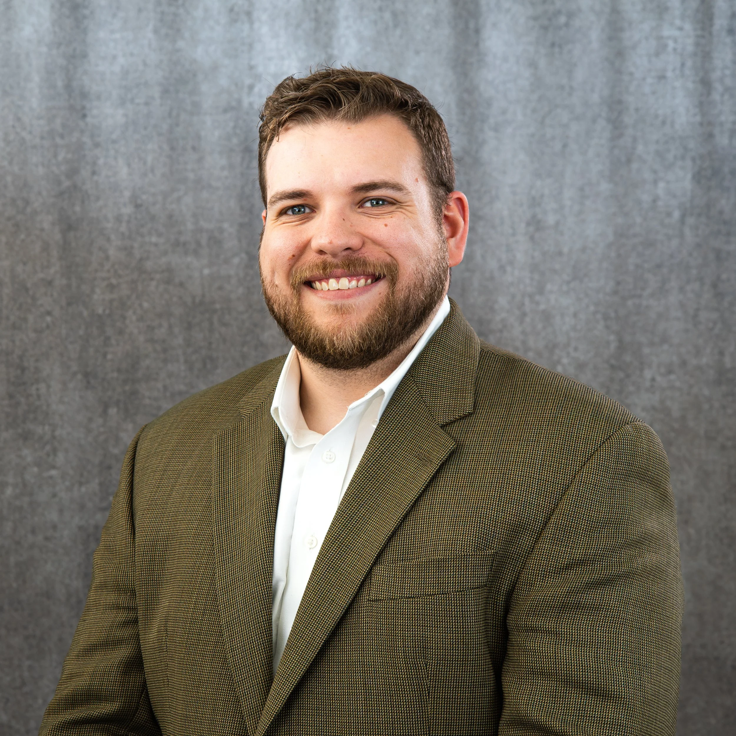 A smiling man with short brown hair, blue eyes, and a beard, wearing a white shirt and a brown blazer, stands against a grey backdrop.
