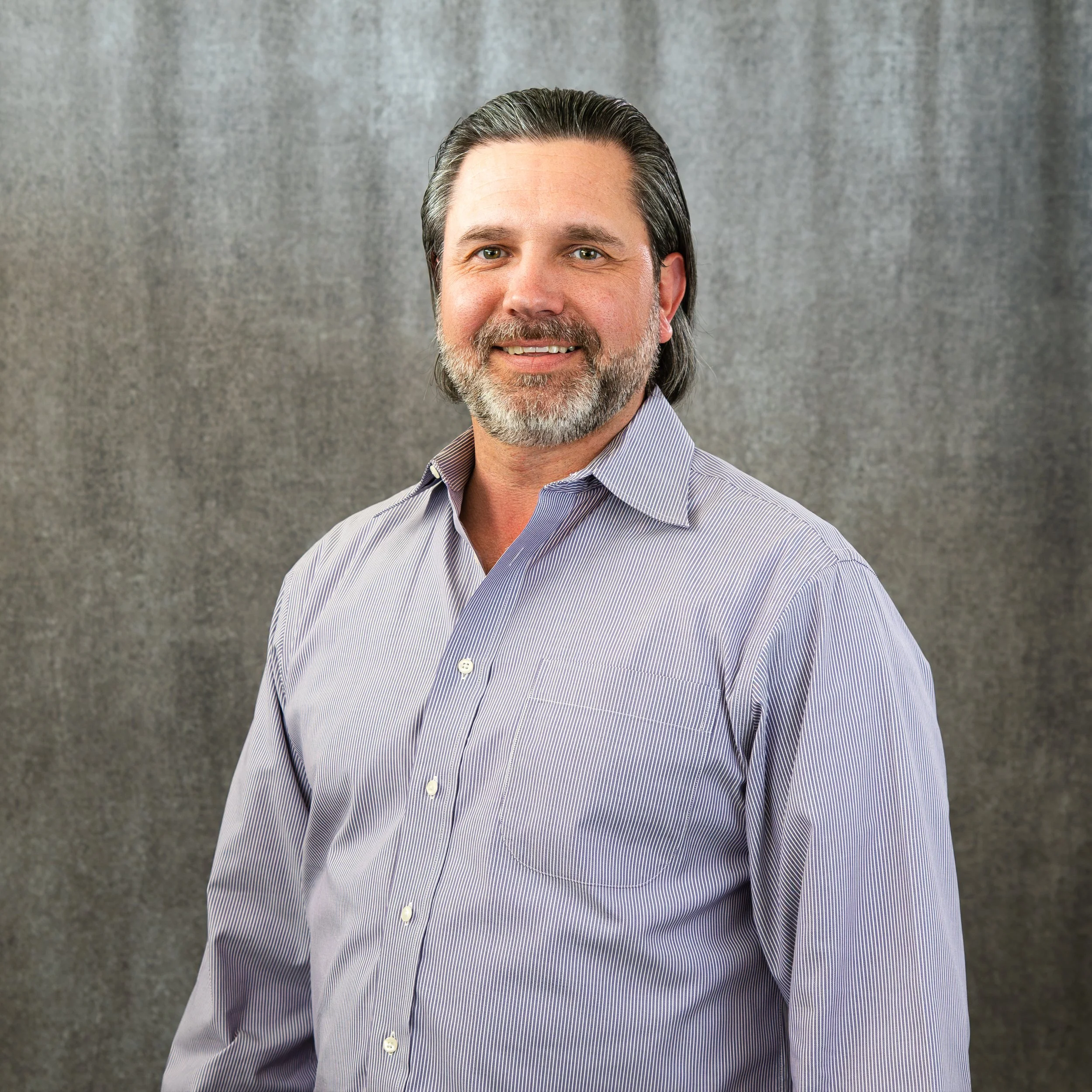 A professional man with gray hair, beard, and mustache wearing a light blue striped dress shirt, smiling in front of a gray textured background.