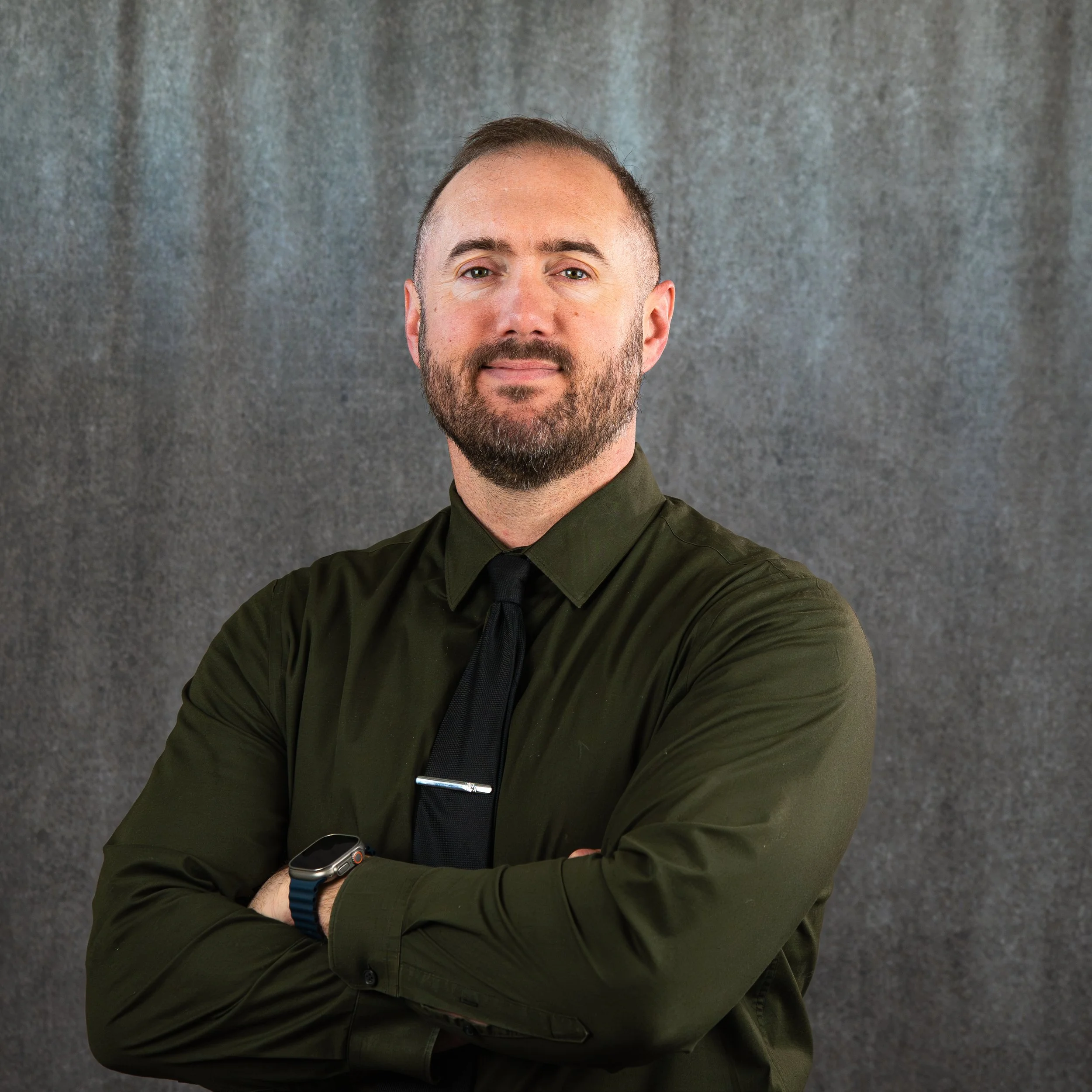 A man with short hair and a beard wearing an olive green shirt, black tie, and smartwatch, crossing his arms in front of a gray textured background.