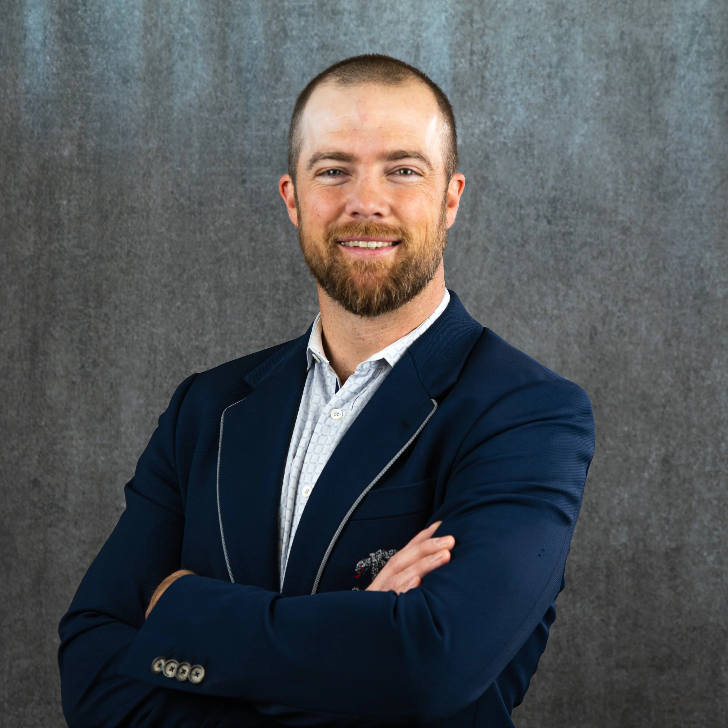 A man in a dark blue blazer and white shirt smiling against a gray textured background.
