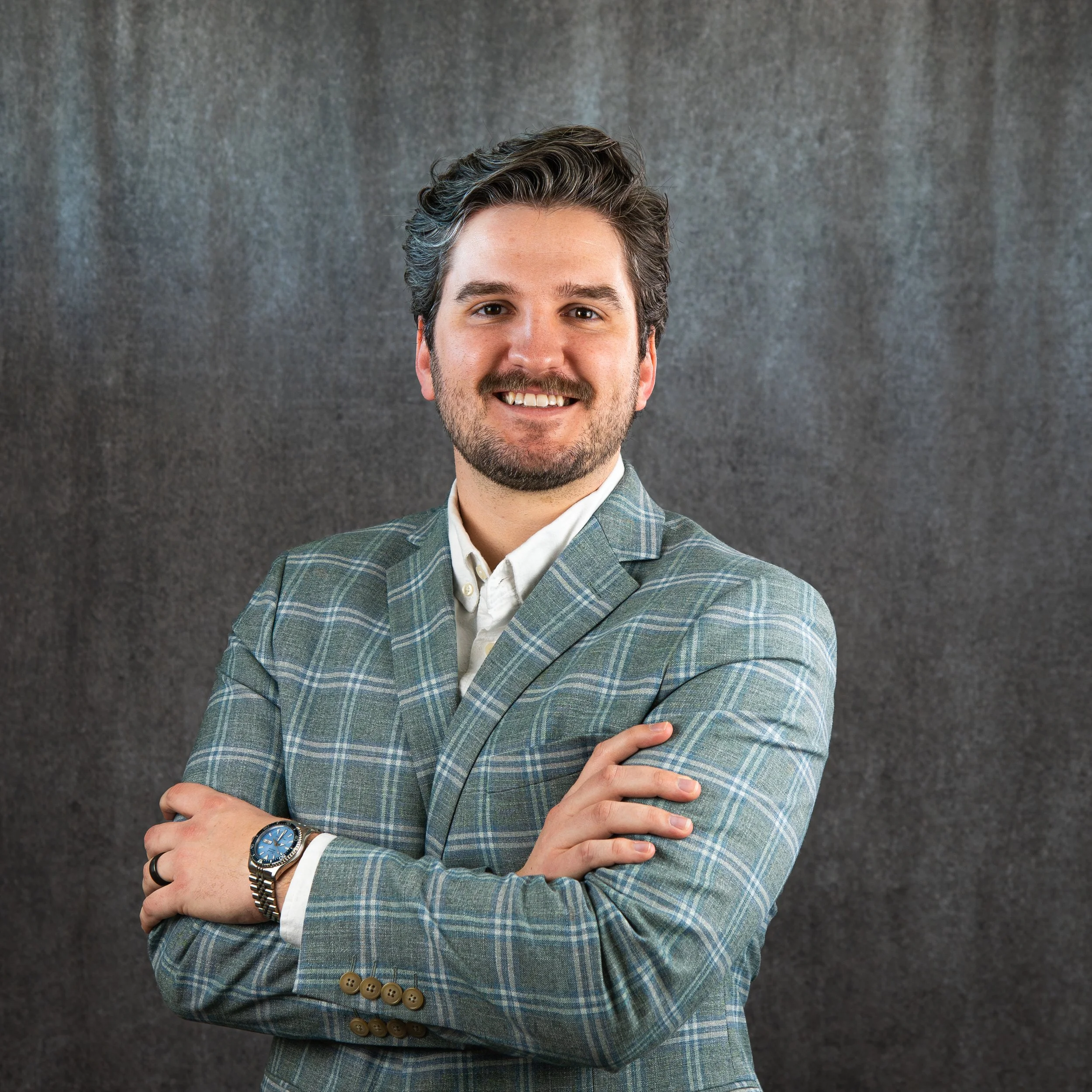 A man with dark, wavy hair and a beard, smiling, dressed in a patterned green blazer, white shirt, wearing a silver wristwatch, standing against a dark gray textured background.