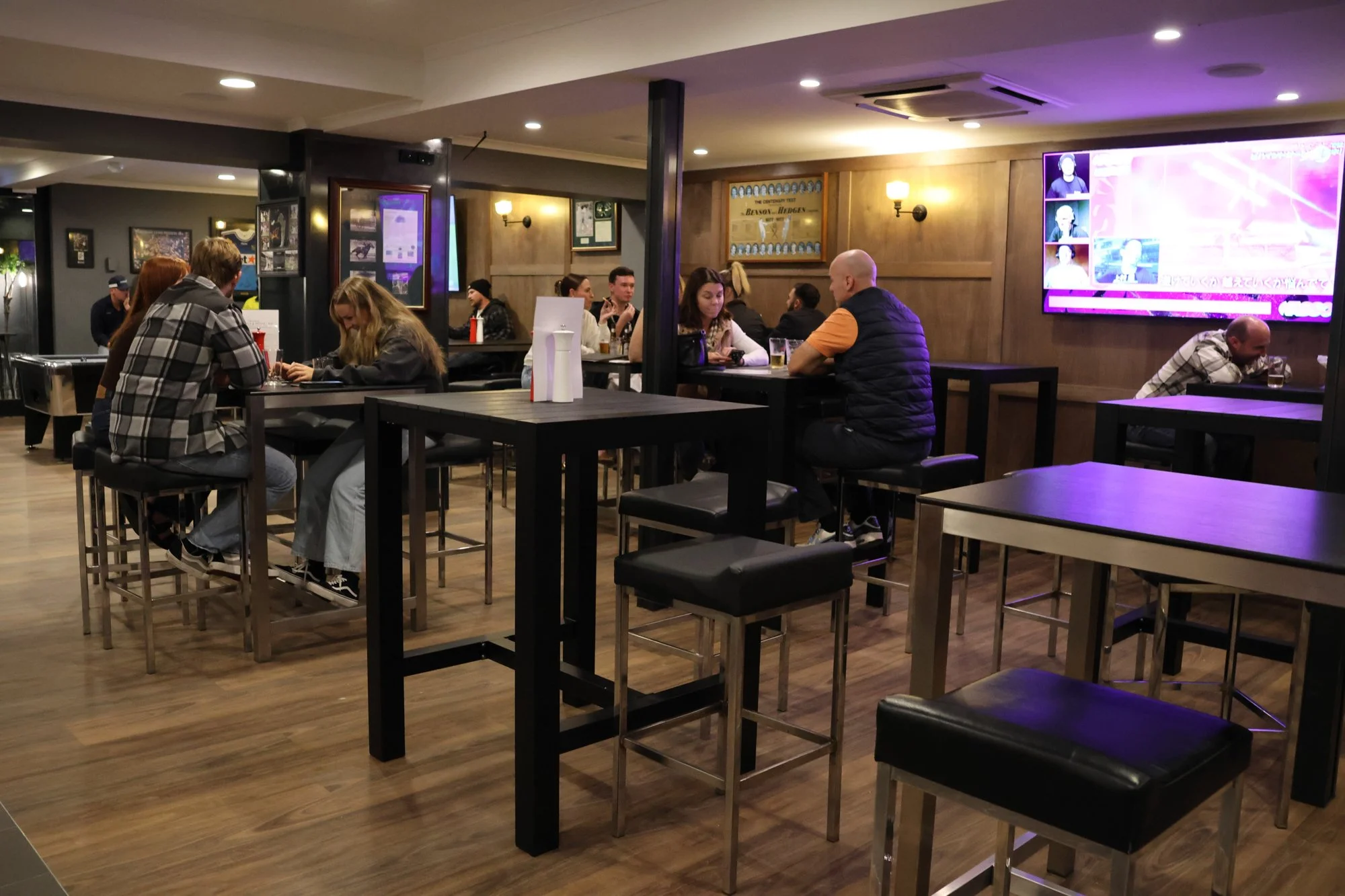 People sitting at high and low tables in a modern bar or pub, watching a TV screen on the wall, with wooden and dark-colored furniture and dim, cozy lighting.