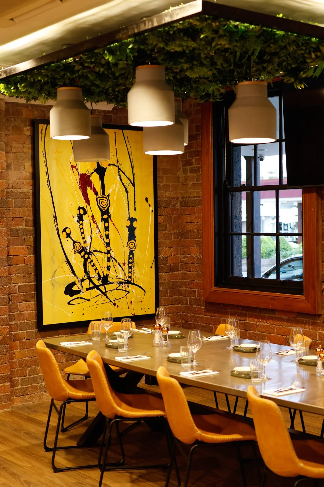 A dining room with a long wooden table set with empty wine glasses and plates, orange chairs, a large abstract yellow painting on a brick wall, and hanging white pendant lights with greenery on the ceiling, alongside a window.