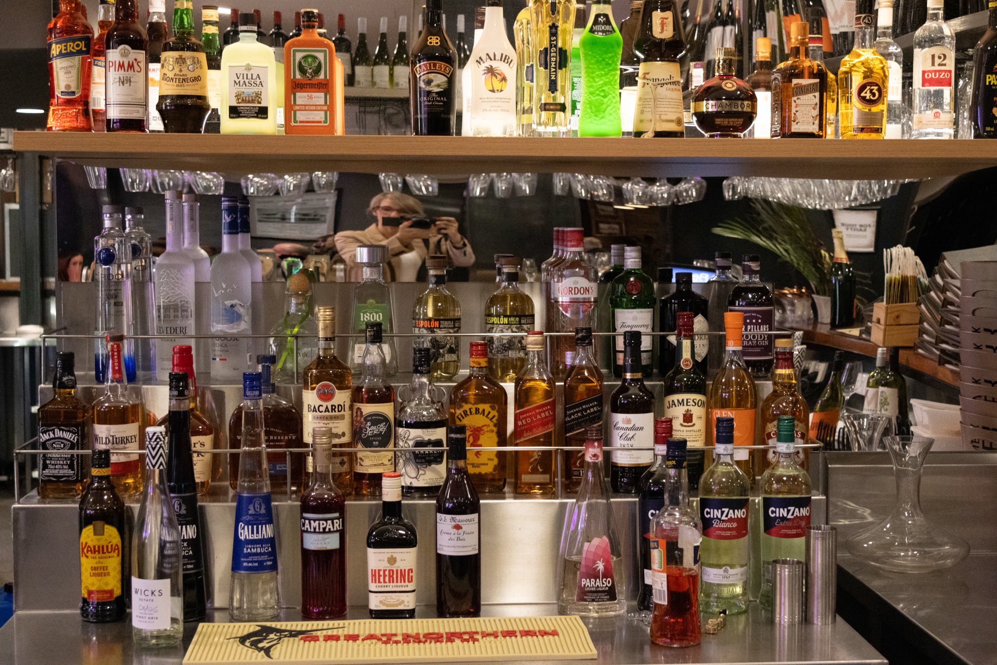 Bar with various bottles of liquor and spirits on glass shelves, including whiskey, vodka, gin, and liqueurs, with a person taking a photo reflected in the mirror behind the bar.
