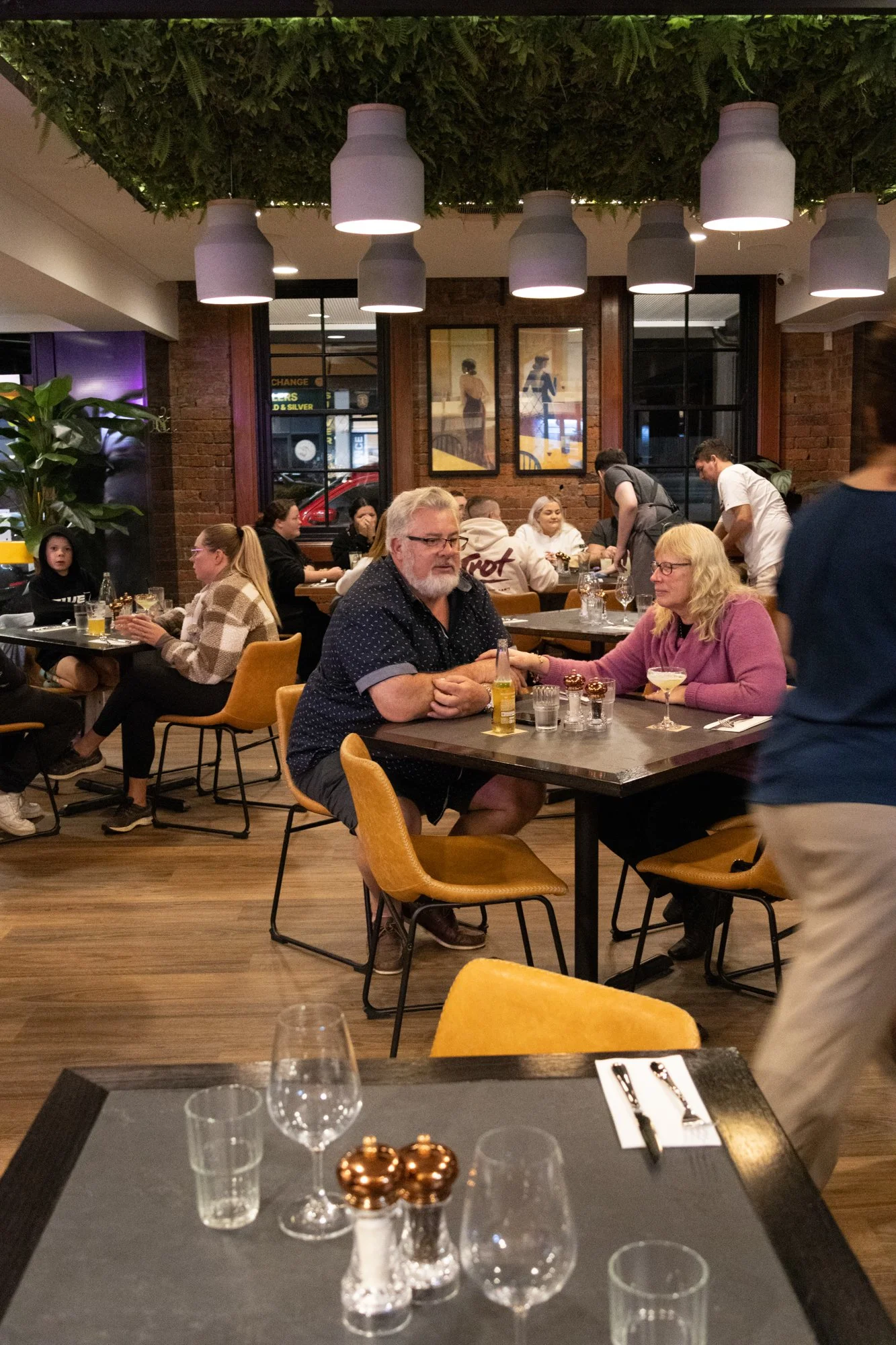 People dining in a modern restaurant with brick walls, framed artwork, and hanging lamps, with some servers attending to tables.