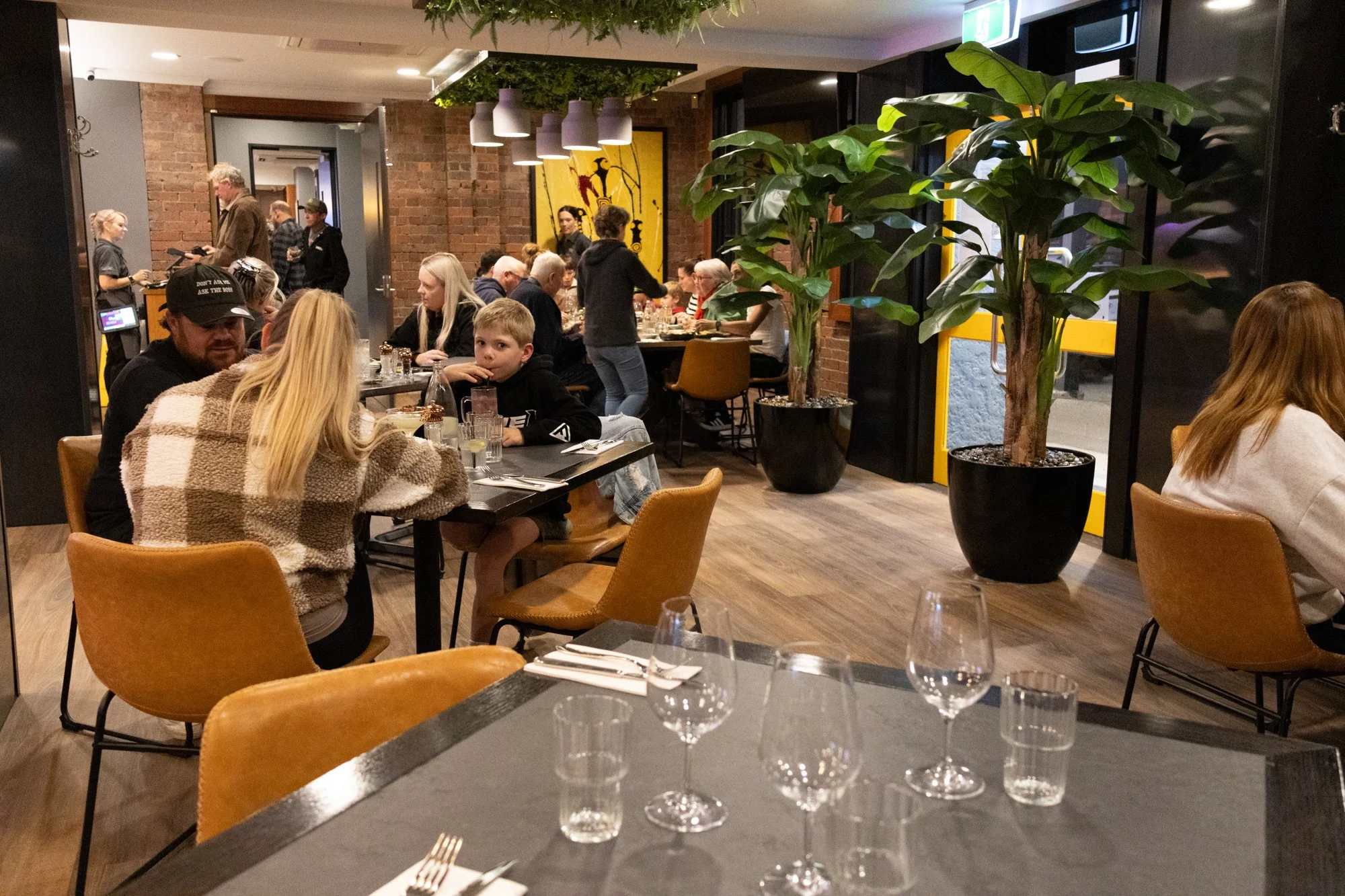 Inside a busy restaurant with people dining at tables, large indoor plants, brick walls, and modern decor.