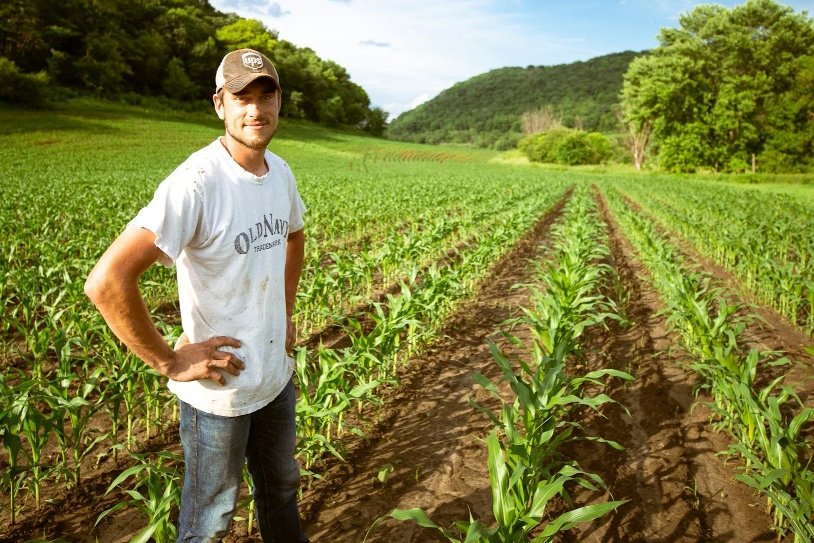 Young farmer wearing t-shirt and hat standing in field of corn