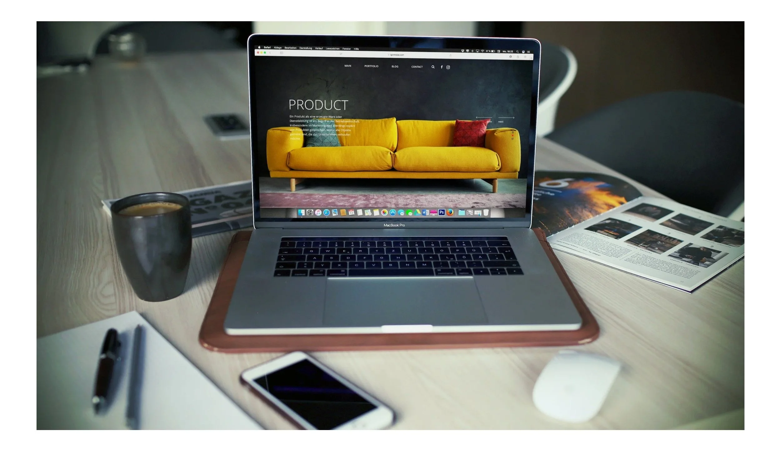 Laptop on a desk displaying a website with a yellow sofa image, with a coffee mug, smartphone, pen, paper, and magazine surrounding it.