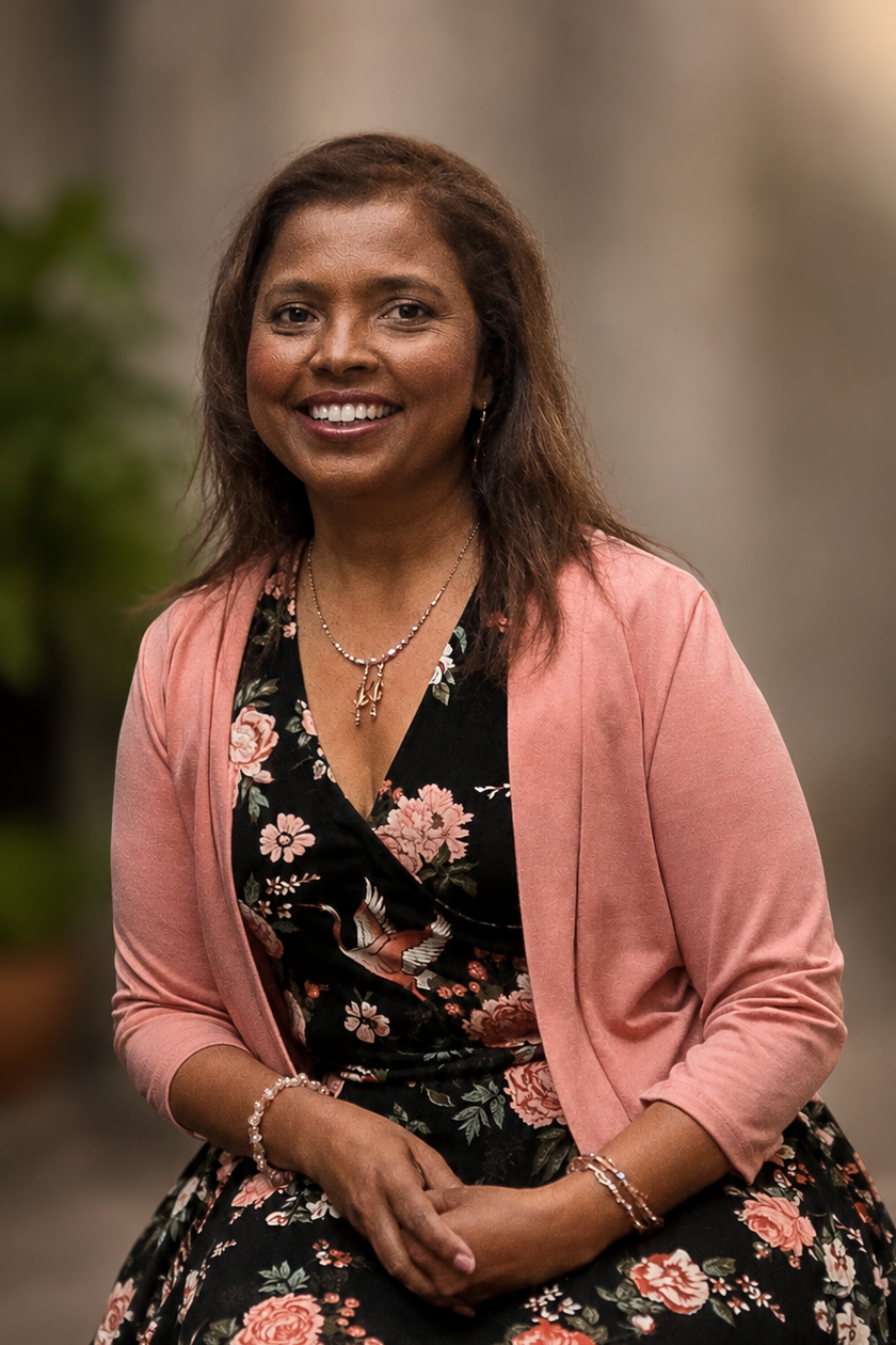 A smiling woman with dark hair, wearing a pink blazer over a black floral dress, and accessorized with necklaces and bracelets, sitting indoors with a blurred background.