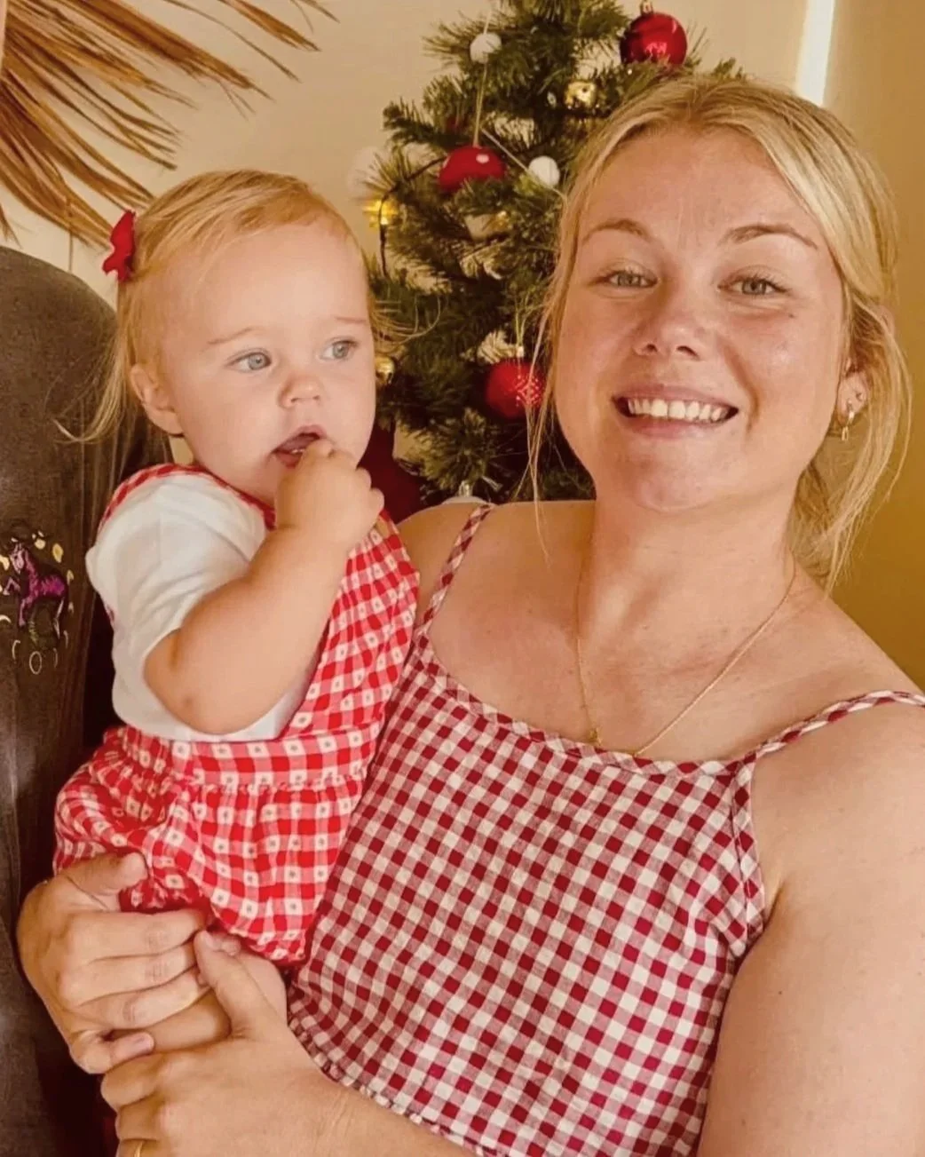 A woman holding a young girl in front of a decorated Christmas tree. The woman is smiling, wearing a red and white checkered dress. The girl has light hair, with a red bow, and is wearing a matching red and white checkered dress, with her finger in her mouth. The background features Christmas ornaments on the tree.