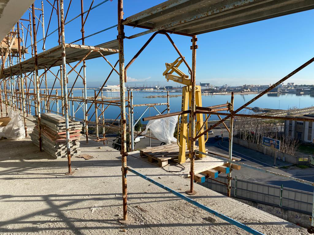 Scaffolding in ferro e legno su un cantiere con vista sul mare, con un cielo azzurro e un porto sullo sfondo.
