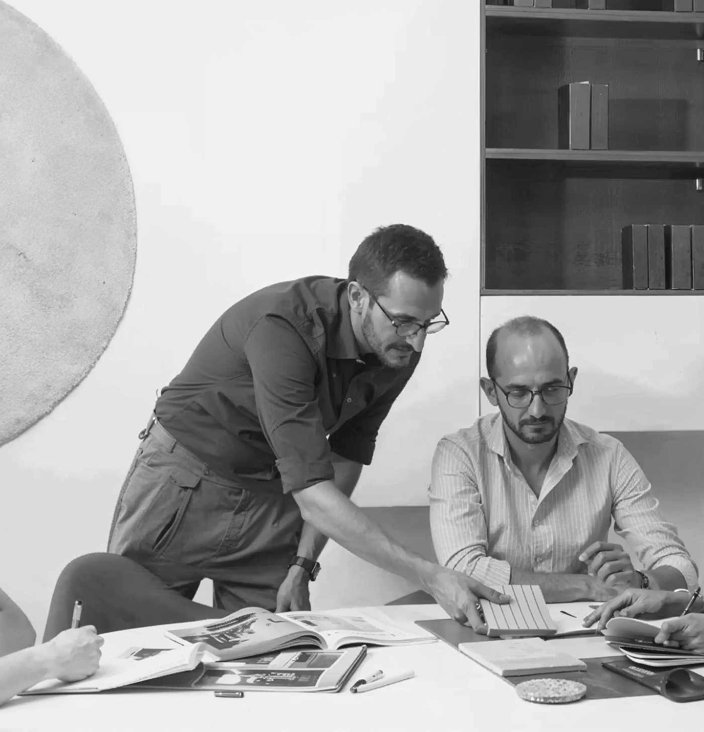 Due uomini con occhiali lavorano insieme su documenti e riviste su un tavolo in un ufficio, uno dei due sta mostrando un quaderno all'altro.