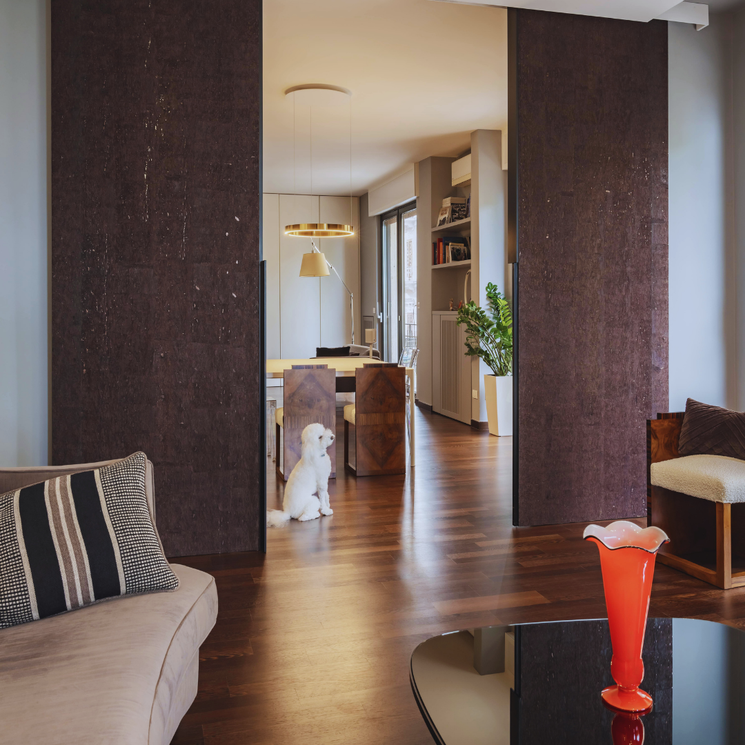 Interno di un soggiorno con mobili in stile moderno, divano beige con cuscino a righe, tavolino nero con una scultura rossa, e una porta scorrevole che apre su una sala da pranzo con pavimenti in legno, una pianta in vaso e una lampada che illumina la zona pranzo.