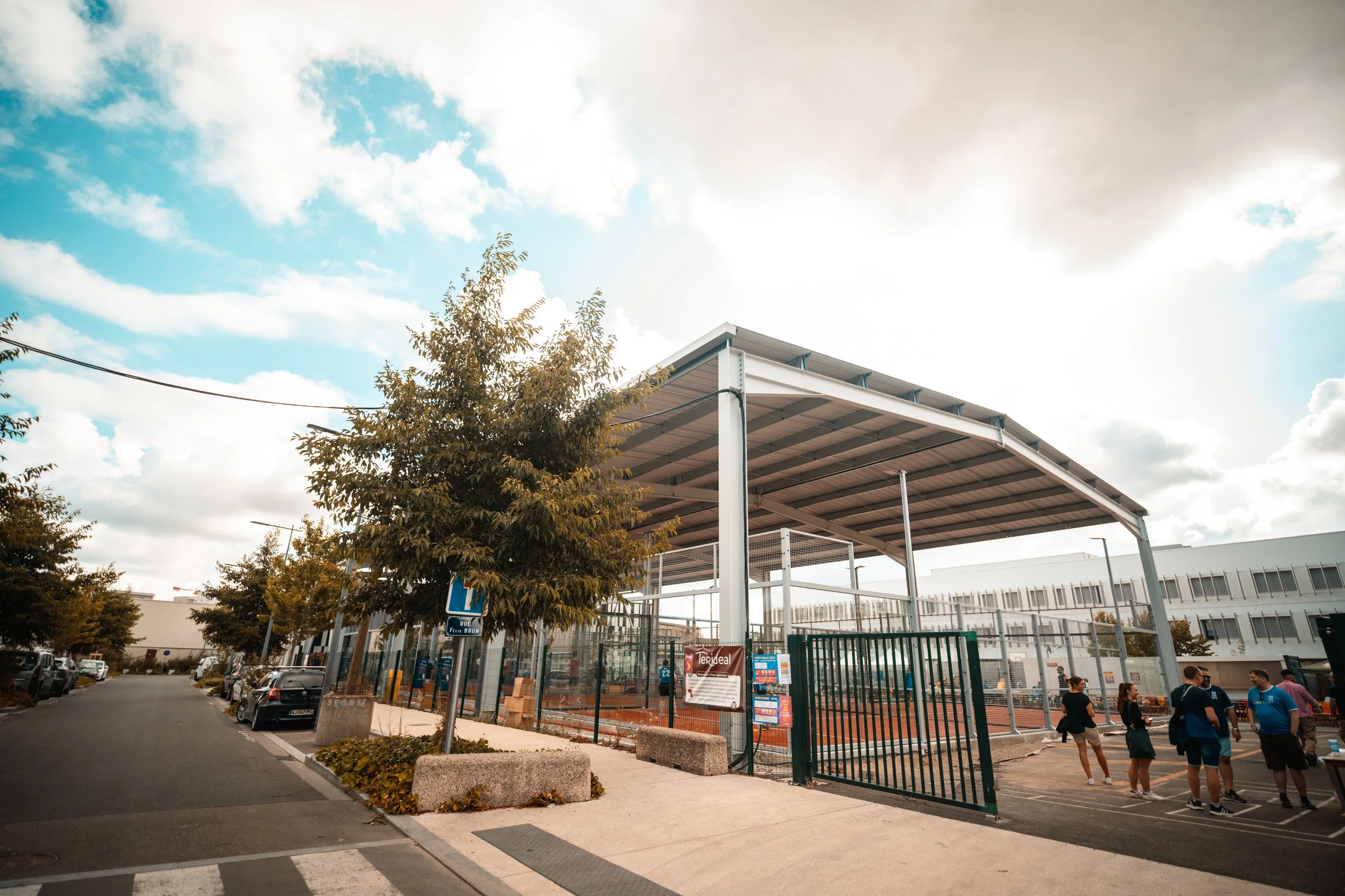 Une aire de sport extérieure avec un toit en métal, entourée de grilles, et des personnes qui attendent ou discutent. Il y a des arbres et des voitures garées le long de la rue.