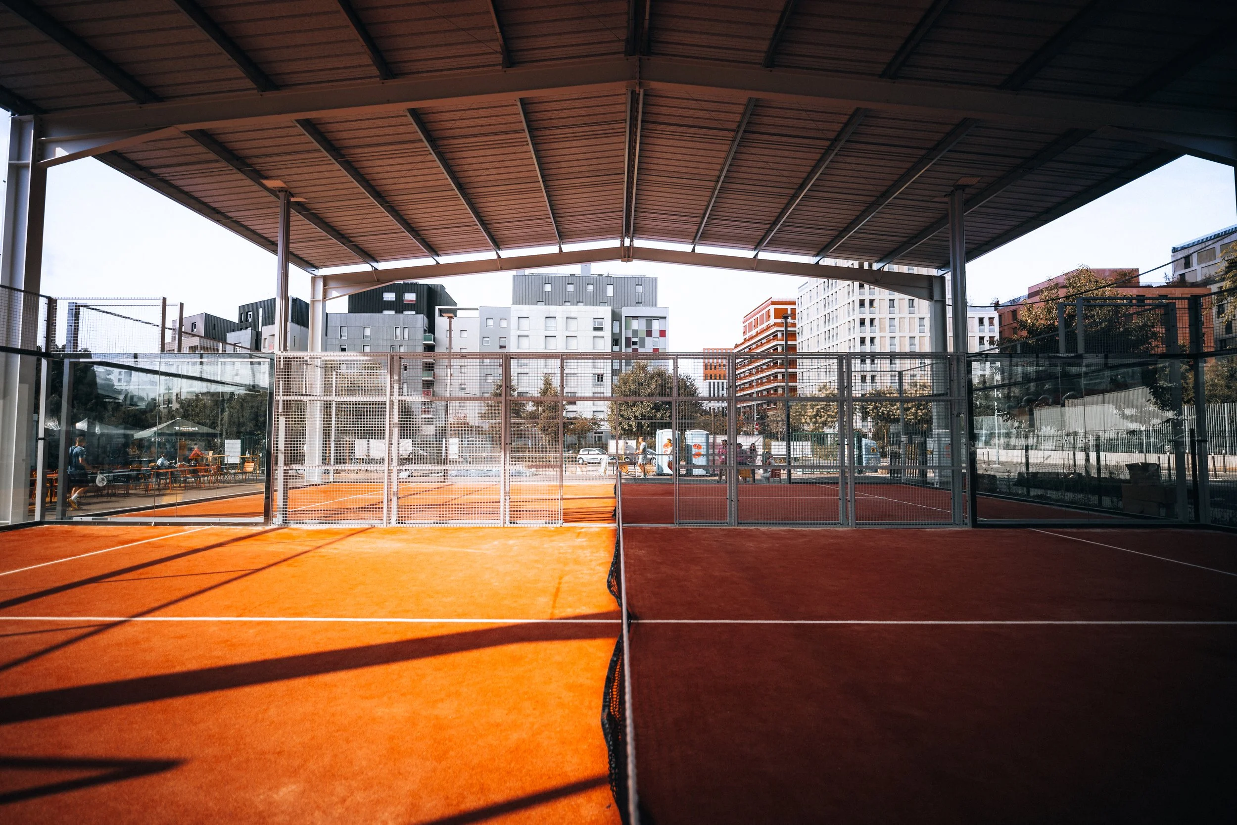 Terrains de padel à lyon en plein air sous un grand toit, entourés de clôtures en verre, avec des bâtiments modernes en arrière-plan.