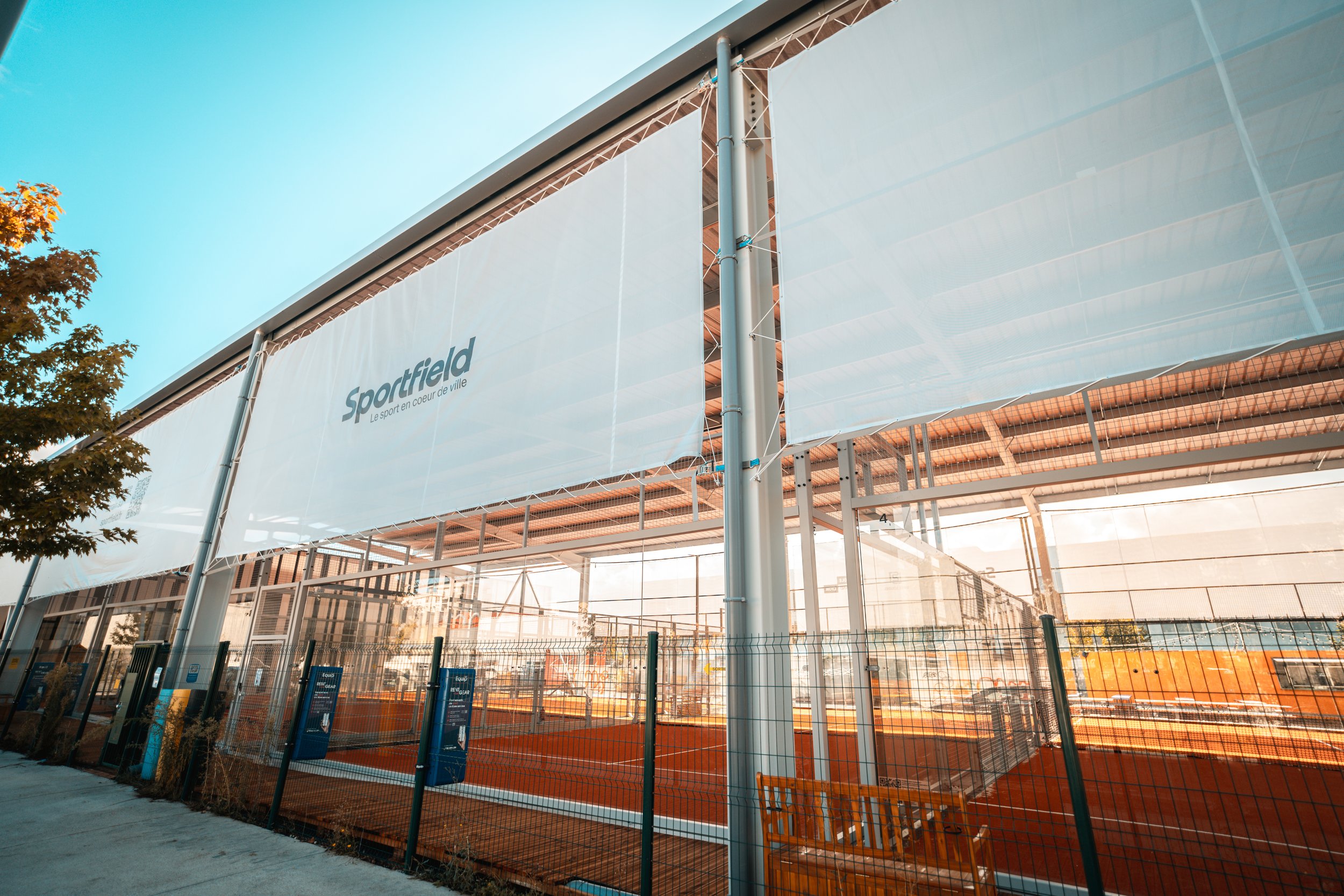 Terrains de padel en plein air entourés d'une clôture, avec une bannière indiquant 'Sportfield' et le slogan 'Le sport en cœur de ville', sous un ciel bleu.