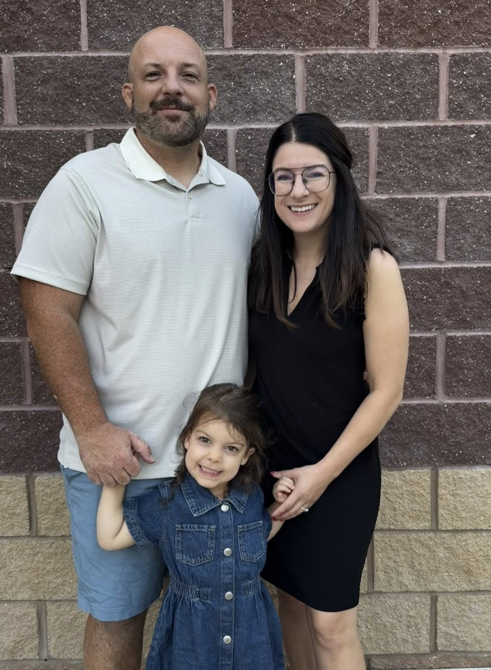 A family of three standing in front of a brick wall. The man has a beard and is wearing a white polo shirt and light blue shorts. The woman has long dark hair, glasses, and is wearing a black sleeveless dress. The young girl has brown hair, a denim dress, and is smiling brightly.