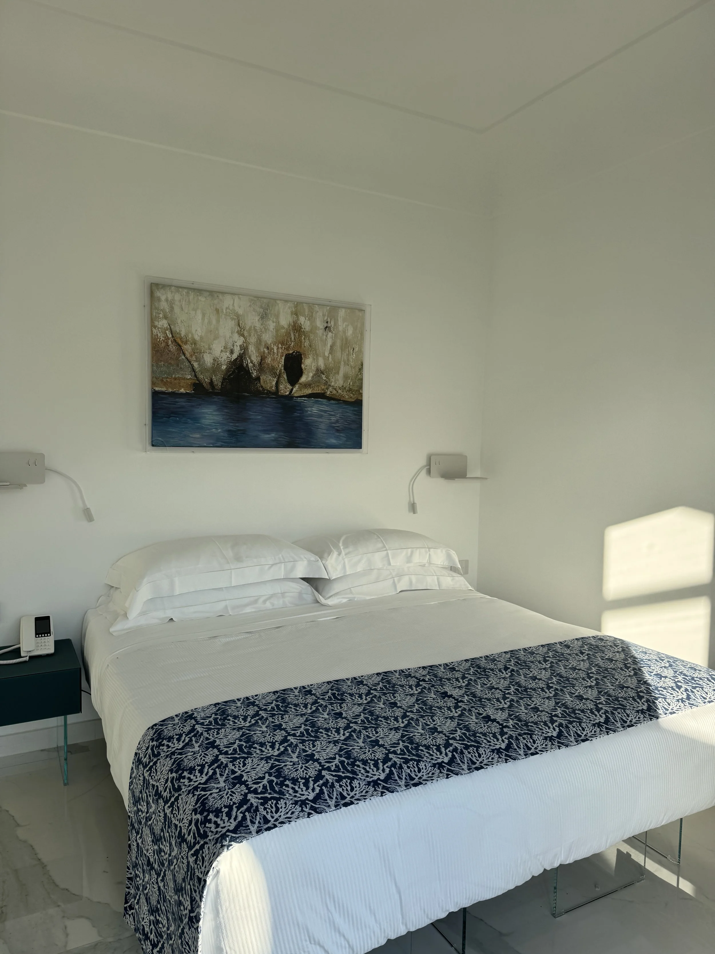 Bright, white bedroom with a bed covered in white and blue pillows and a blue bedspread with a coral pattern, glass shower enclosure in the background, glass door leading outside, and a wall-mounted flat-screen TV.