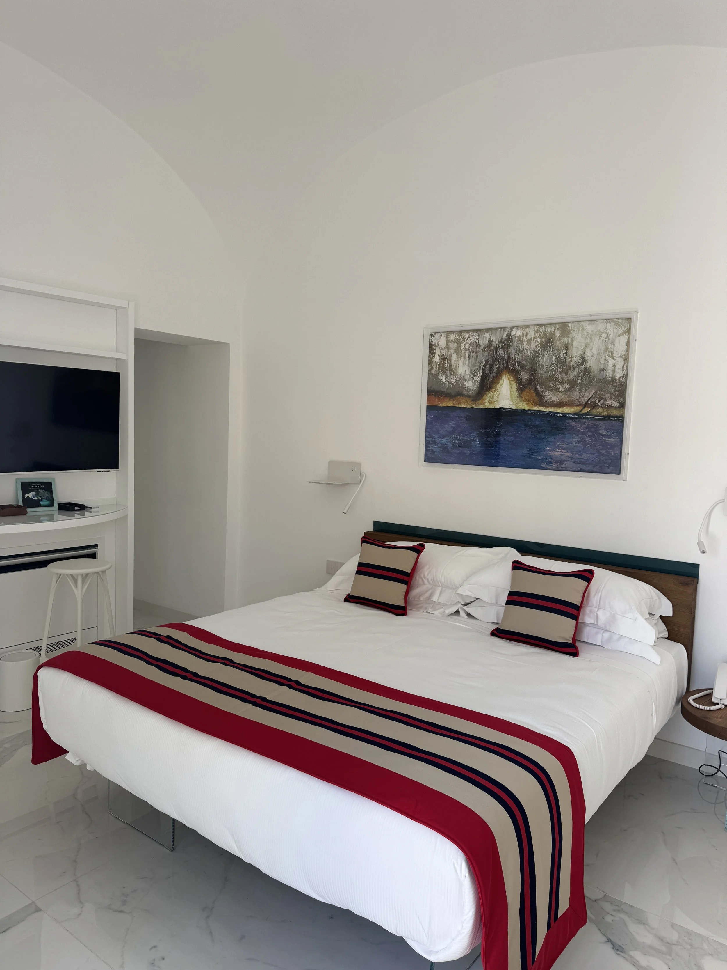 Bright bedroom with white walls and marble floor, large sliding glass doors leading to a balcony, a wall-mounted TV, a bed with white bedding and blue patterned pillows,