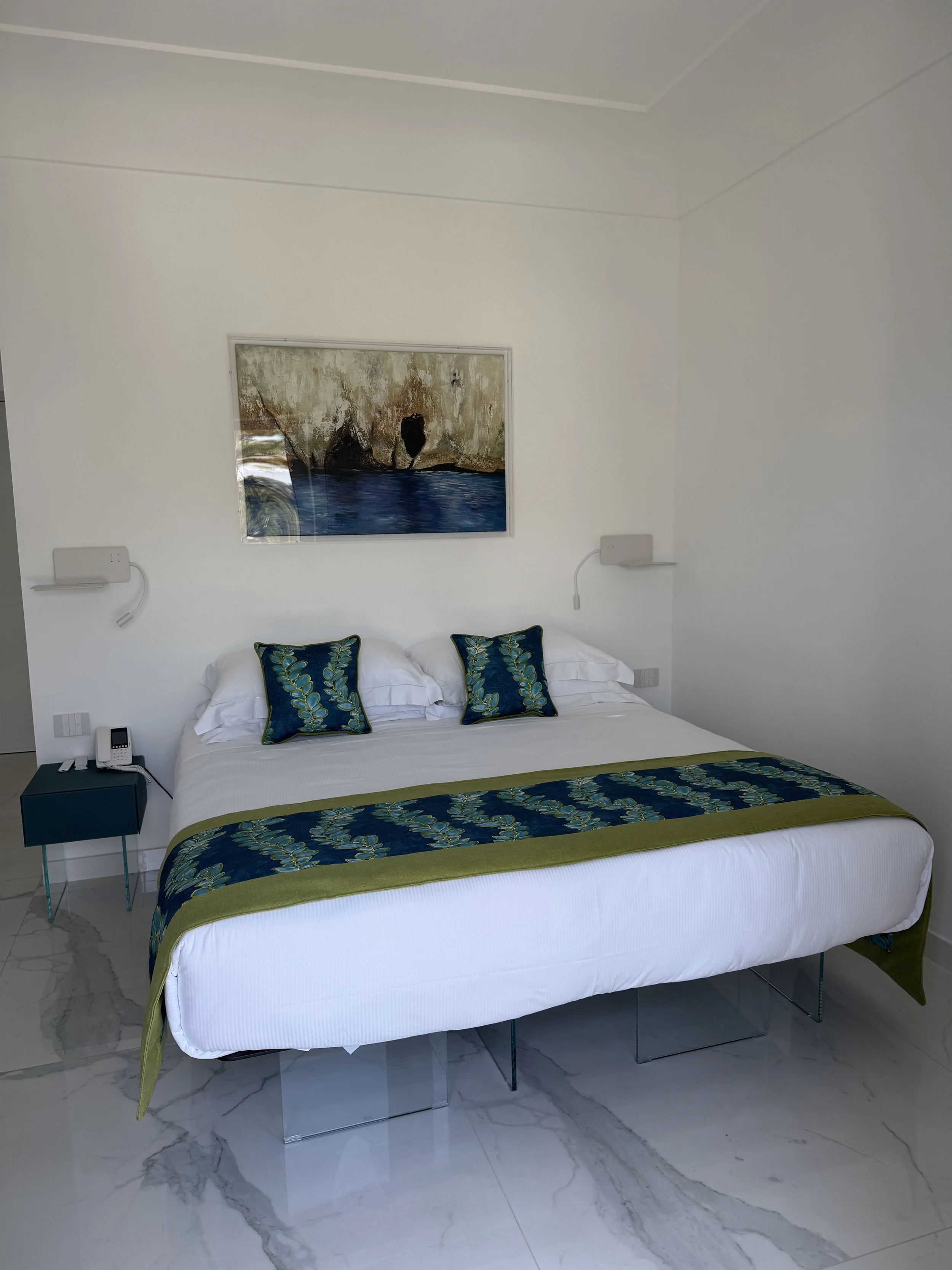 Bright, white bedroom with a bed covered in white and blue pillows and a blue bedspread with a coral pattern, glass shower enclosure in the background, glass door leading outside, and a wall-mounted flat-screen TV.