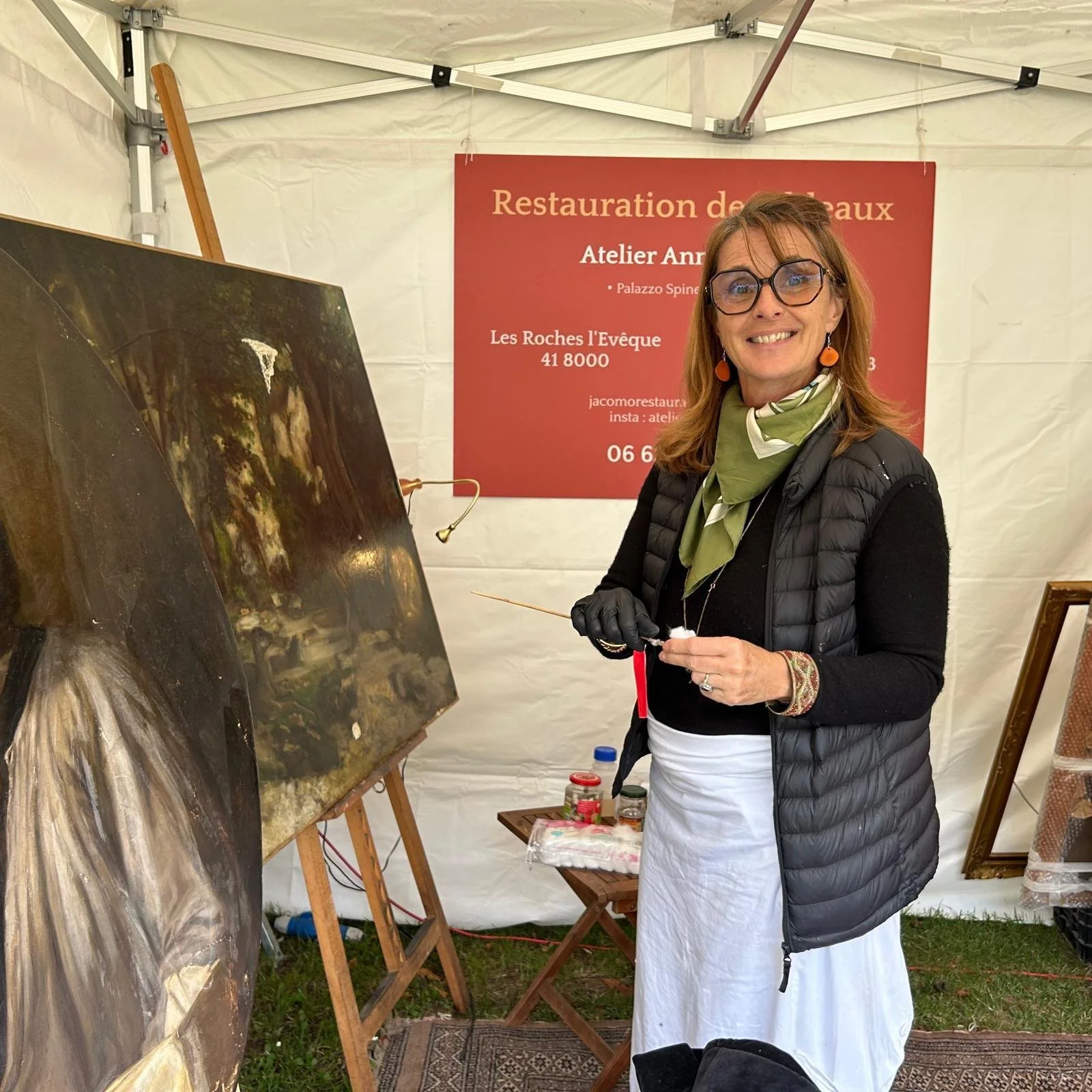 Femme peignant une toile lors d'un événement artistique en extérieur, portant un manteau noir, un foulard vert, des lunettes, et un tablier blanc, avec un panneau rouge en arrière-plan.