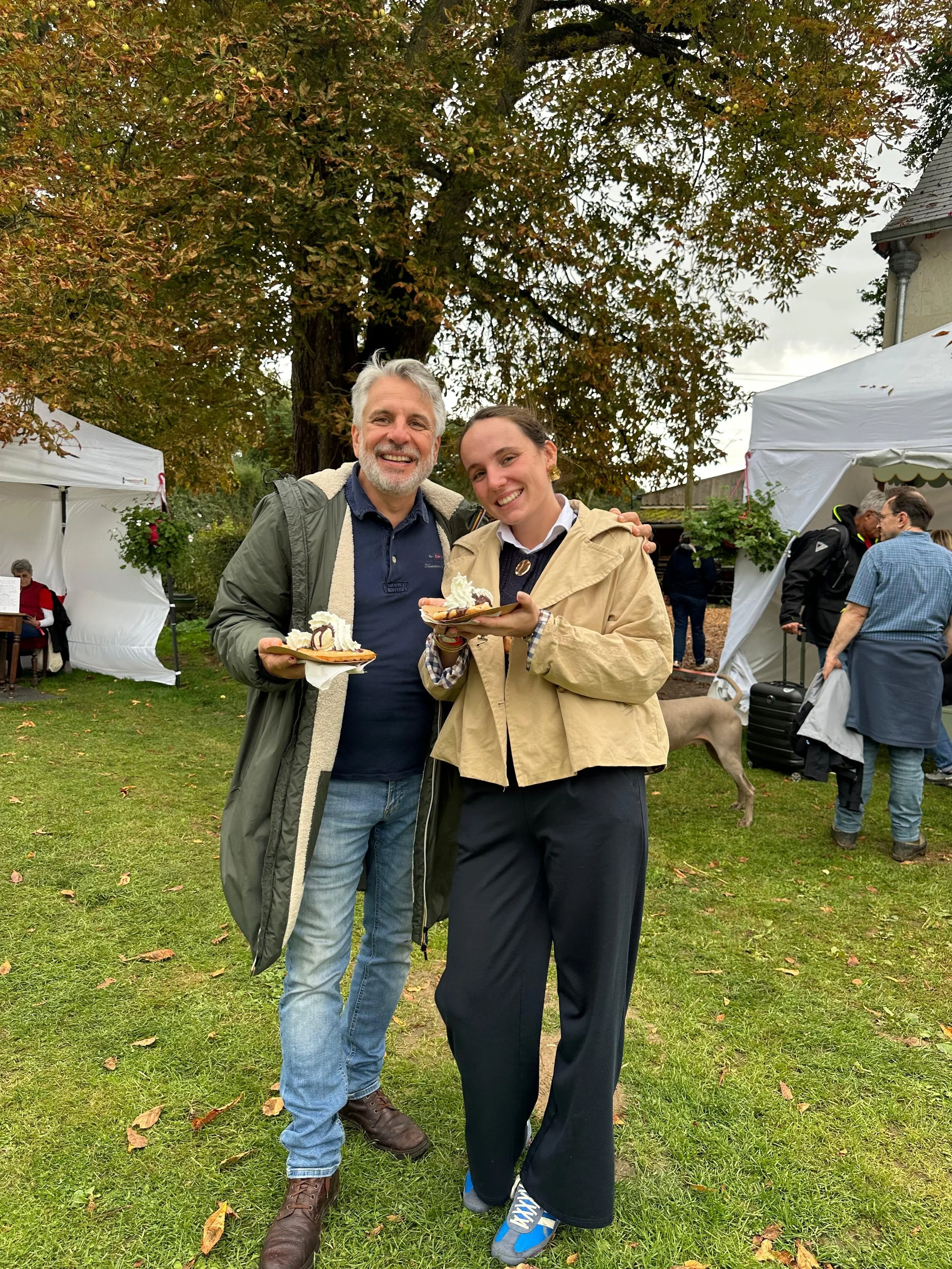 Deux personnes souriantes tenant une assiette avec une pâtisserie, debout sur une pelouse lors d'un événement en plein air, avec des tentes blanches et des arbres en arrière-plan.