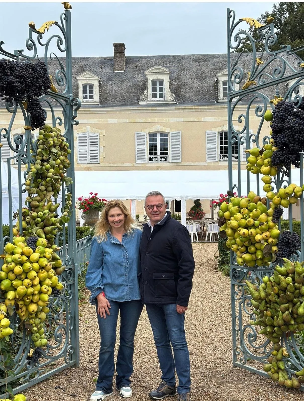 Deux personnes posent sous une arche de vines en métal orné de grappes de raisins, devant un grand bâtiment avec fenêtres à volets et un toit en ardoise.