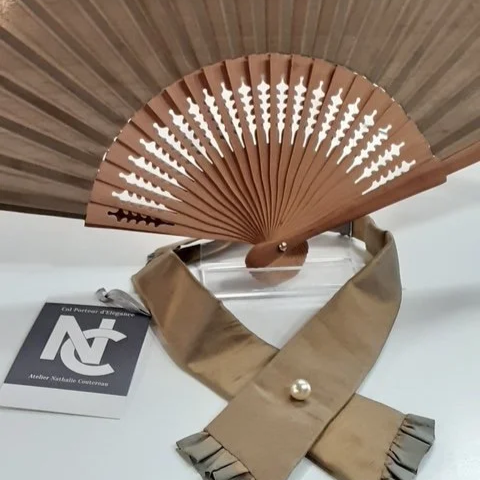 Éventail en bois plié avec motif décoratif, posé sur un sac beige avec une perle, à côté d'une étiquette de l'Inde.