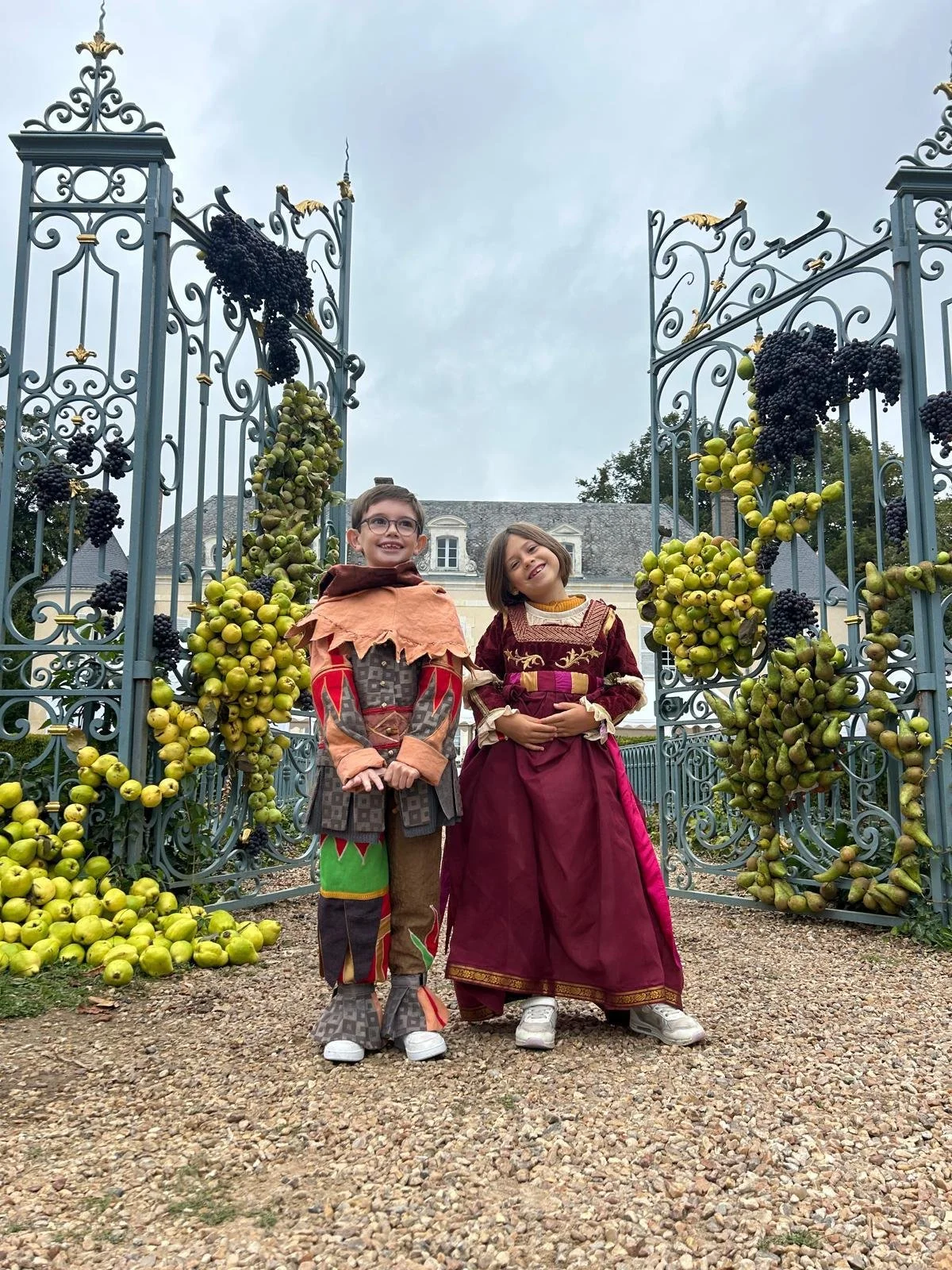 Deux enfants habillés en costumes médiévaux, un garçon et une fille, se tiennent devant une porte en fer ornée de grappes de raisins et de pommes, avec un château ou une grande maison en arrière-plan.