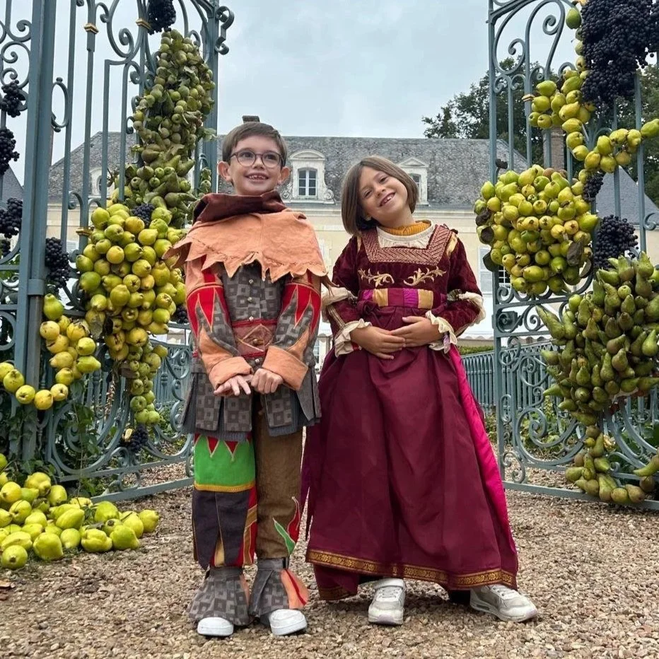 Deux enfants déguisés en personnages médiévaux ou fantastiques, posant sous une arche décorée de grappes de raisins verts et noirs, dans une cour devant un bâtiment ancien.