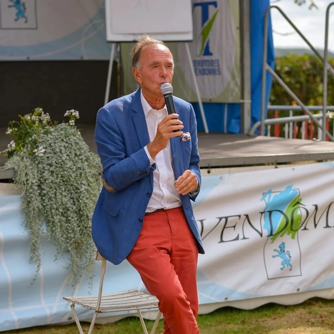 Homme en costume bleu et pantalon rouge, parlant dans un microphone lors d'un événement en plein air.