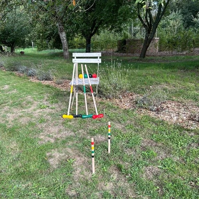Un jeu d'extérieur pour enfants avec une cible en forme de paniers percés, une raquette, deux bâtons colorés devant une haie et des arbres dans un jardin.