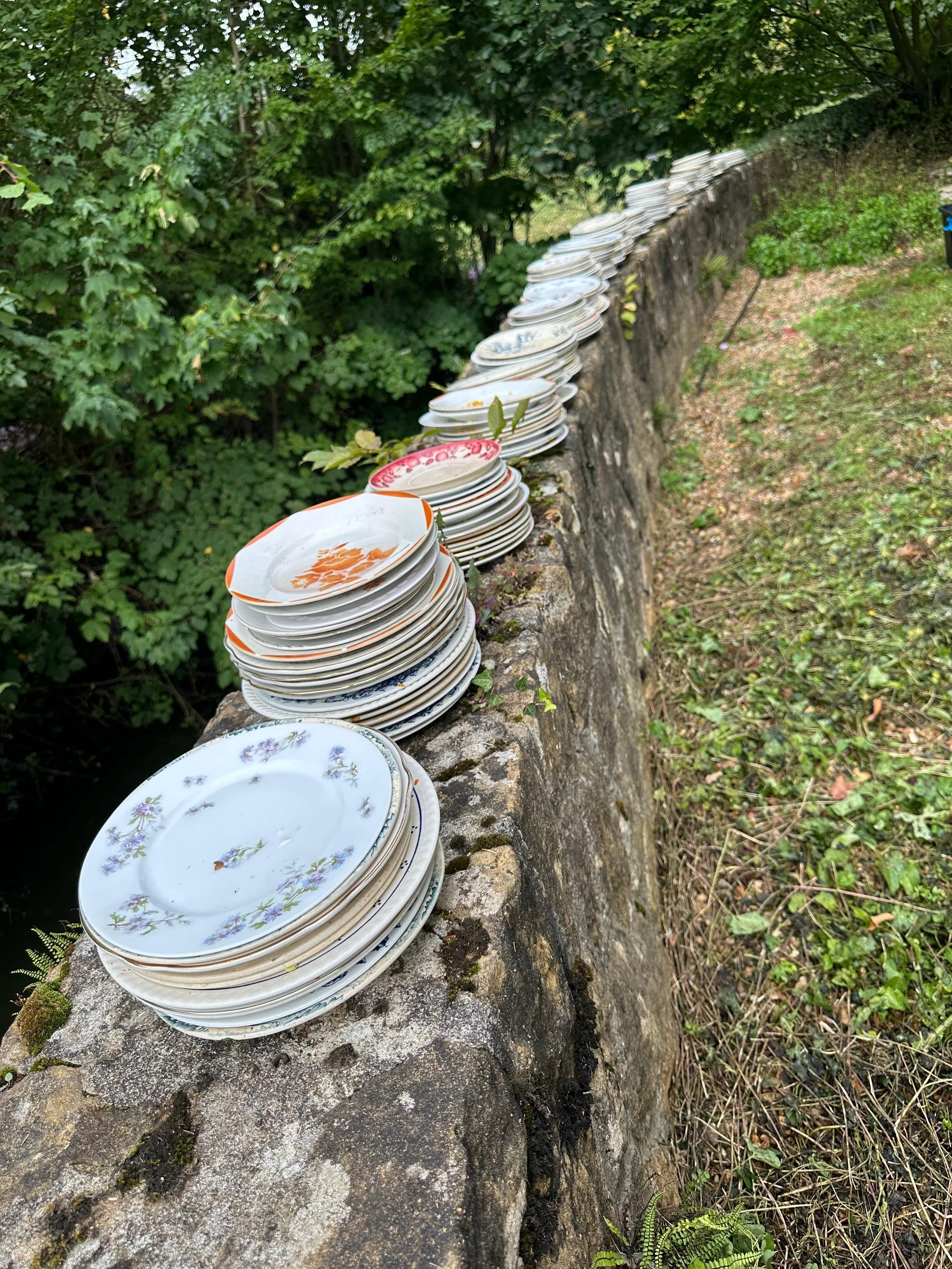 Grand tas de vaisselle en porcelaine avec des motifs floraux, empilée sur un mur en pierre dans un jardin vert.