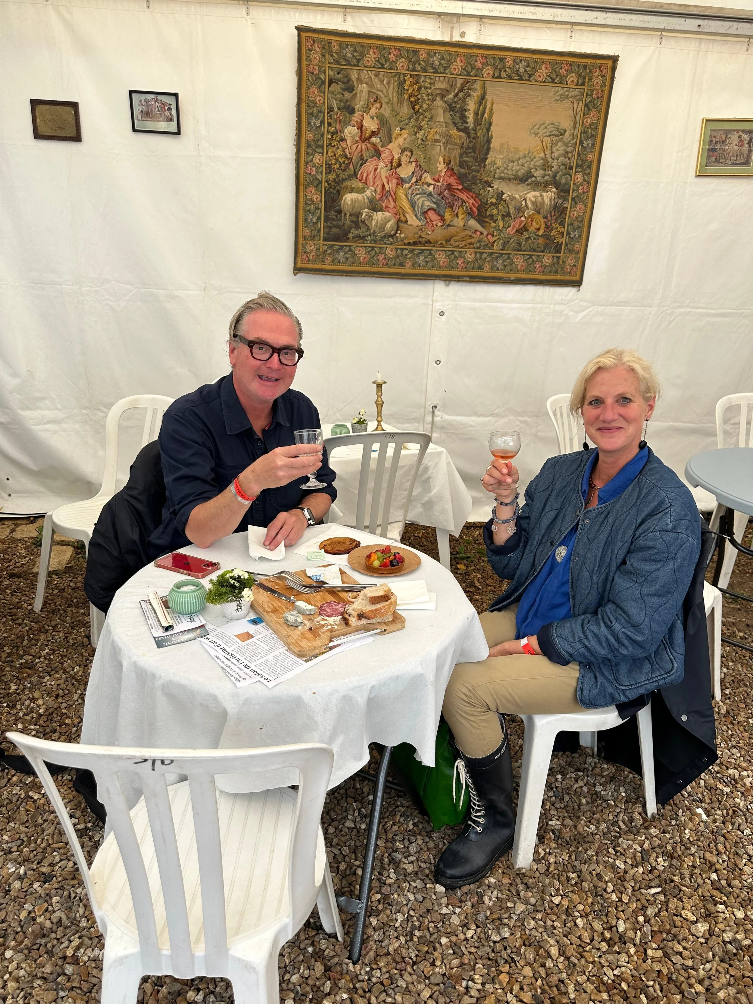 Deux personnes assises à une table ronde en dégustant du vin, avec un plateau de fromages, de la charcuterie et des fruits, dans un cadre intérieur avec un tapis mural et des chaises en plastique.