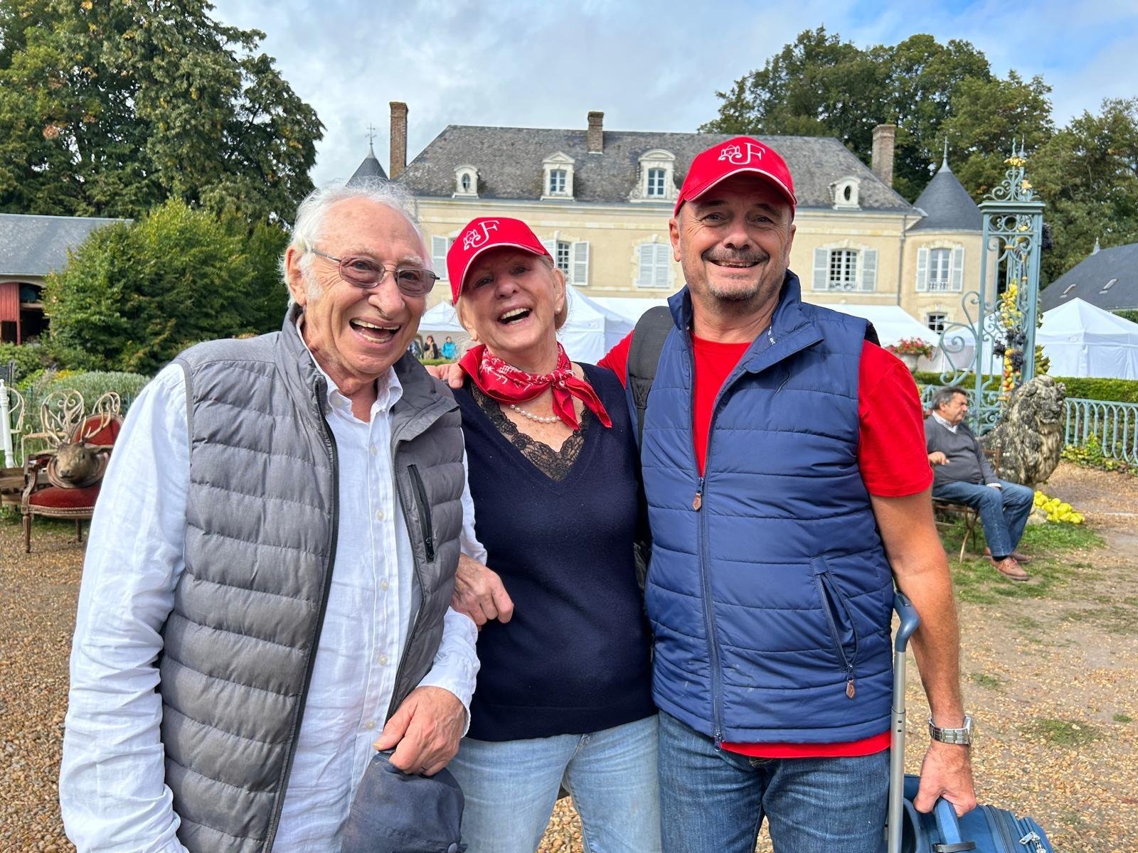 Trois personnes souriantes posent pour une photo à l'extérieur devant un grand bâtiment, deux hommes et une femme, portant tous des vêtements décontractés. La femme et un homme portent des casquettes rouges.